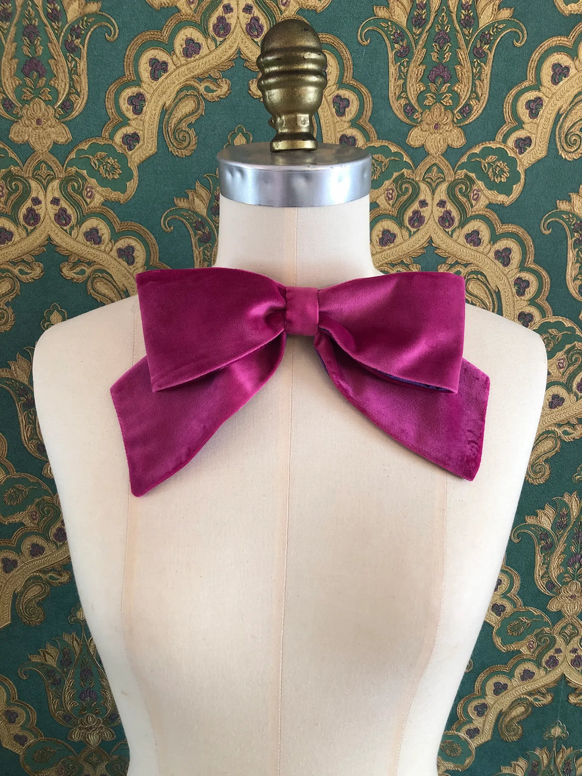 Broochella handmade Large Velvet Bow Brooch in rich magenta velvet with contrasting purple edging, styled asymmetrically on a cream mannequin against ornate green and gold wallpaper.