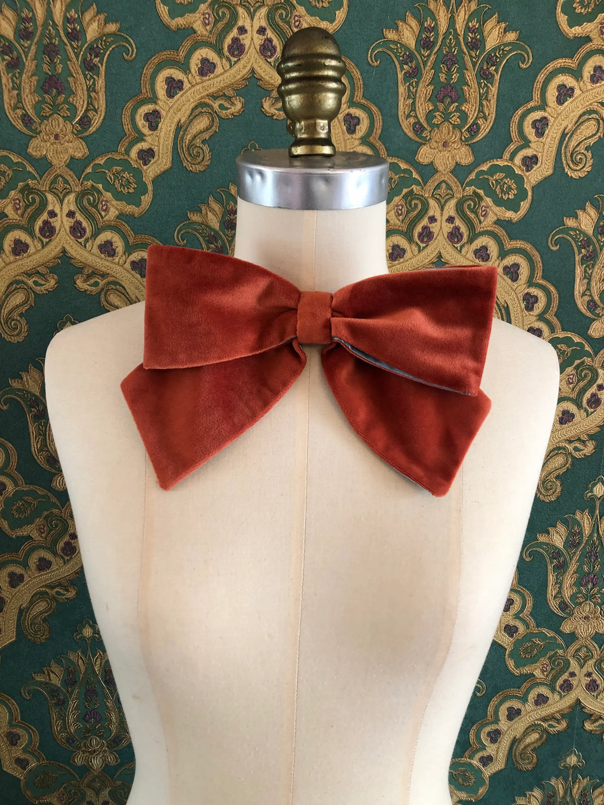Broochella handmade Large Velvet Bow Brooch in rich burnt orange velvet with contrasting ice blue edging, styled asymmetrically on a cream mannequin against ornate green and gold wallpaper.
