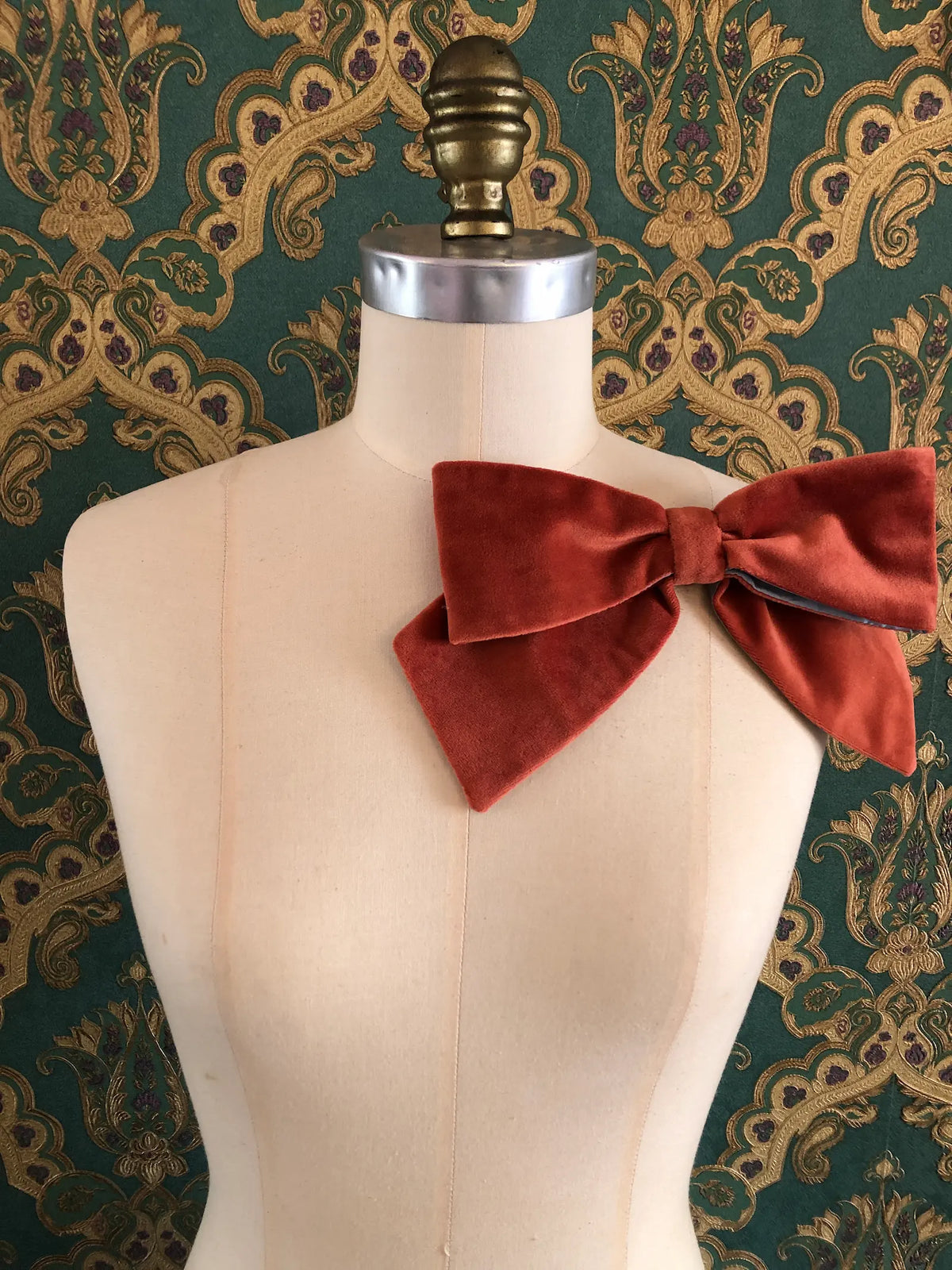 Broochella handmade Large Velvet Bow Brooch in rich burnt orange velvet with contrasting ice blue edging, styled asymmetrically on a cream mannequin against ornate green and gold wallpaper.