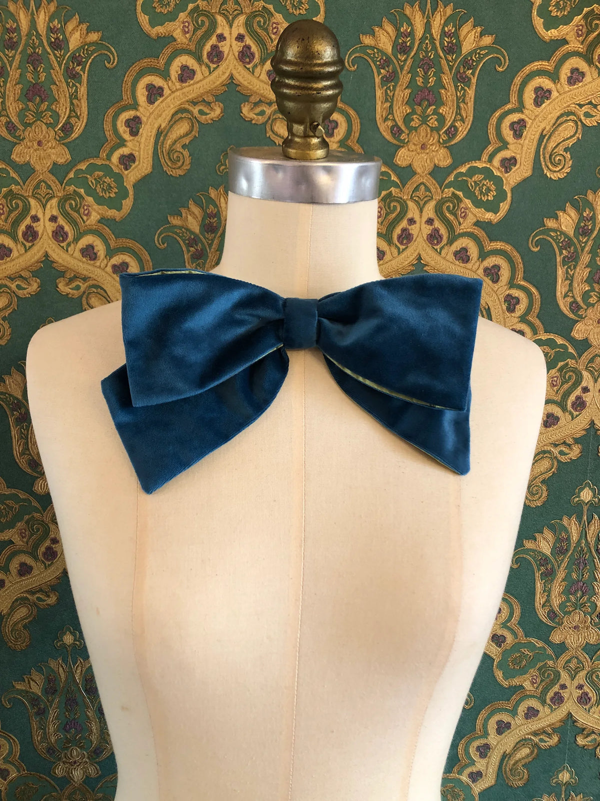 Broochella handmade Large Velvet Bow Brooch in rich peacock blue velvet with contrasting lime green lining, styled asymmetrically on a cream mannequin against ornate green and gold wallpaper.