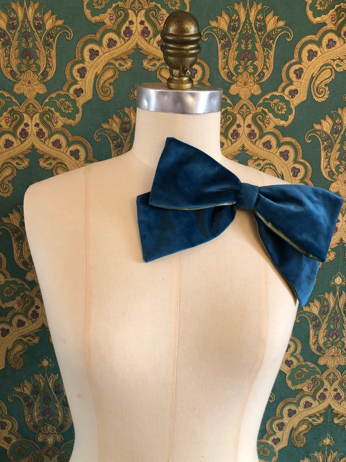 Broochella handmade Large Velvet Bow Brooch in rich peacock blue velvet with contrasting lime green lining, styled asymmetrically on a cream mannequin against ornate green and gold wallpaper.