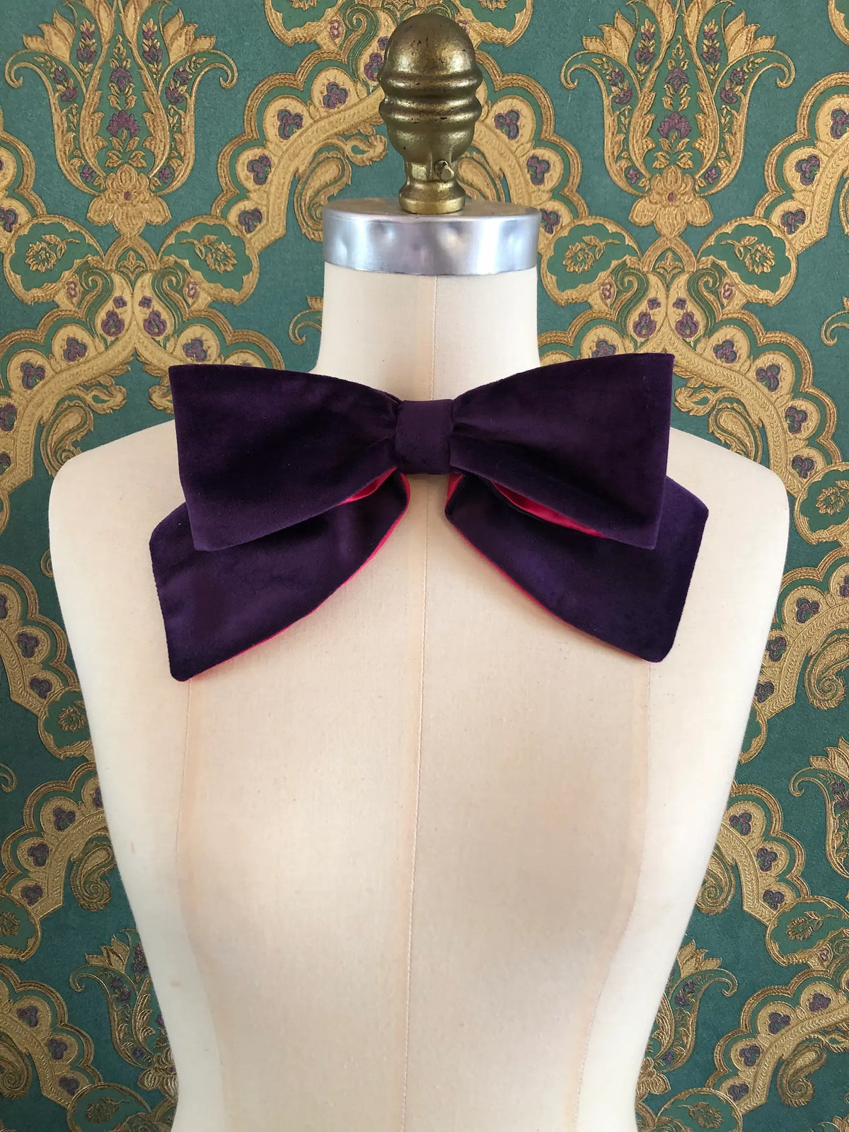 Broochella handmade Large Velvet Bow Brooch in deep purple velvet with contrasting fuchsia lining, styled asymmetrically on a cream mannequin against ornate green and gold wallpaper.