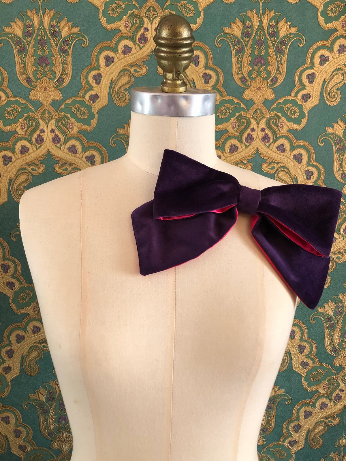 Broochella handmade Large Velvet Bow Brooch in deep purple velvet with contrasting fuchsia lining, styled asymmetrically on a cream mannequin against ornate green and gold wallpaper.