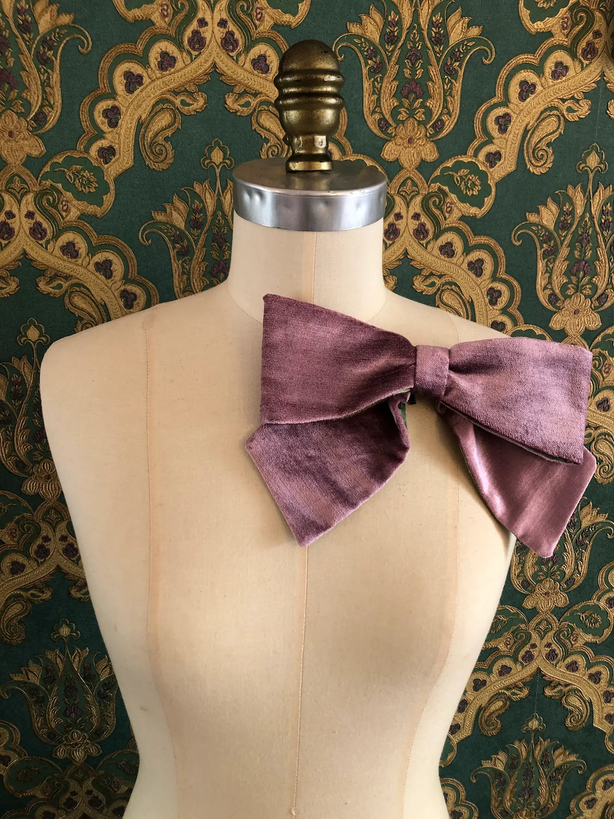 Broochella handmade Large Velvet Bow Brooch in soft violet velvet with contrasting green lining, styled asymmetrically on a cream mannequin against ornate green and gold wallpaper.