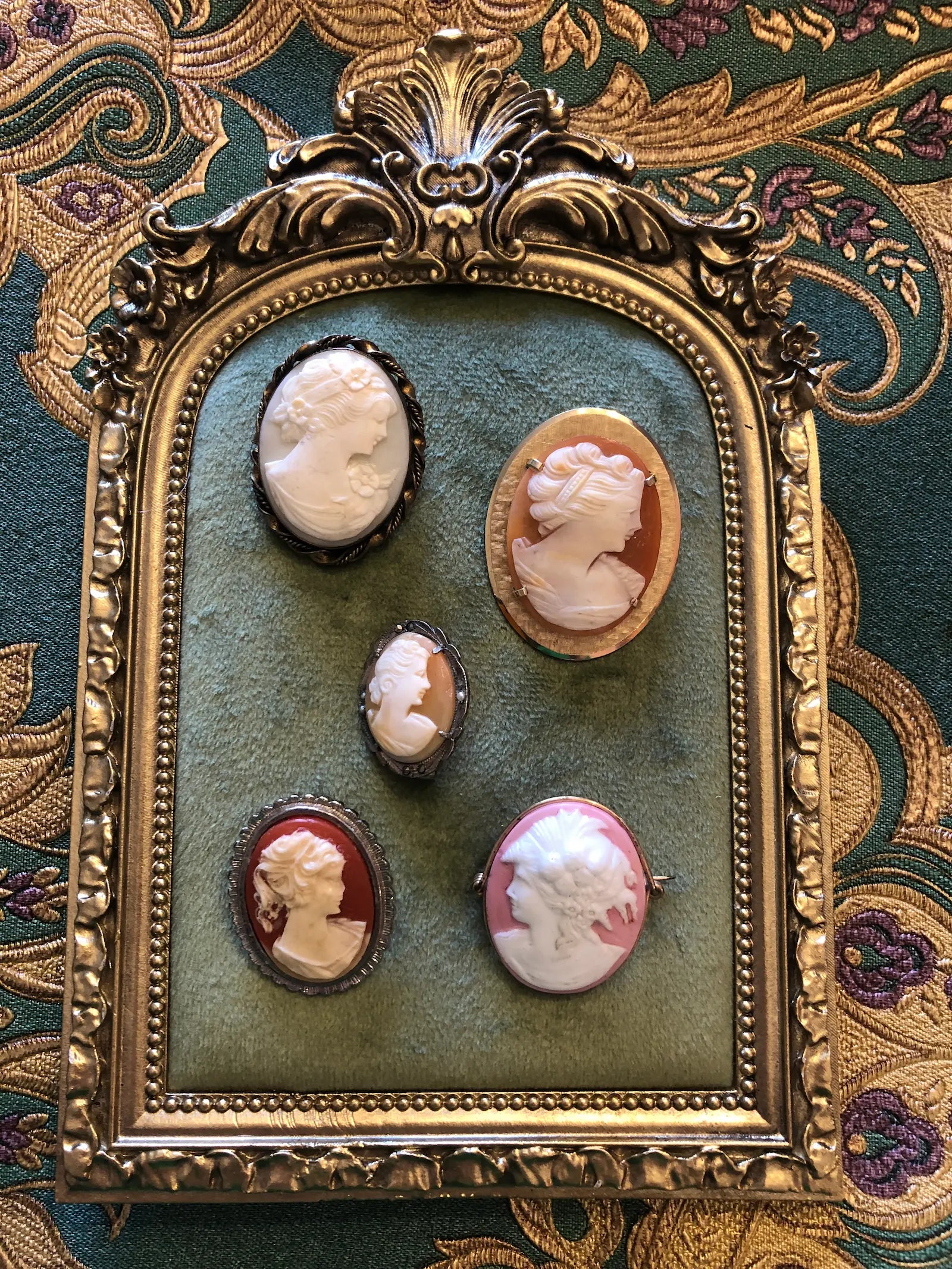 A vintage-style framed brooch display featuring Broochella's le camee five carved cameo-style jewelry pieces in pink, red, and cream tones, mounted on an ornate gold frame over a green fabric backing, set against a patterned textile background.