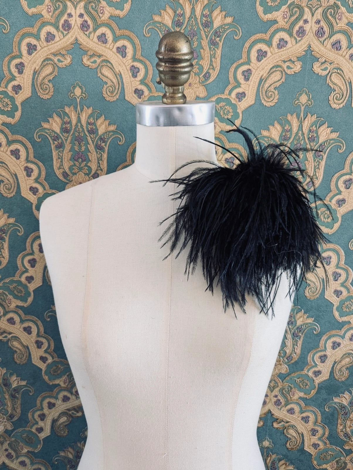 Broochella le pouffe handmade designer feather brooch in black.  It is pinned to a mannequin. The background is green and gold damask wallpaper