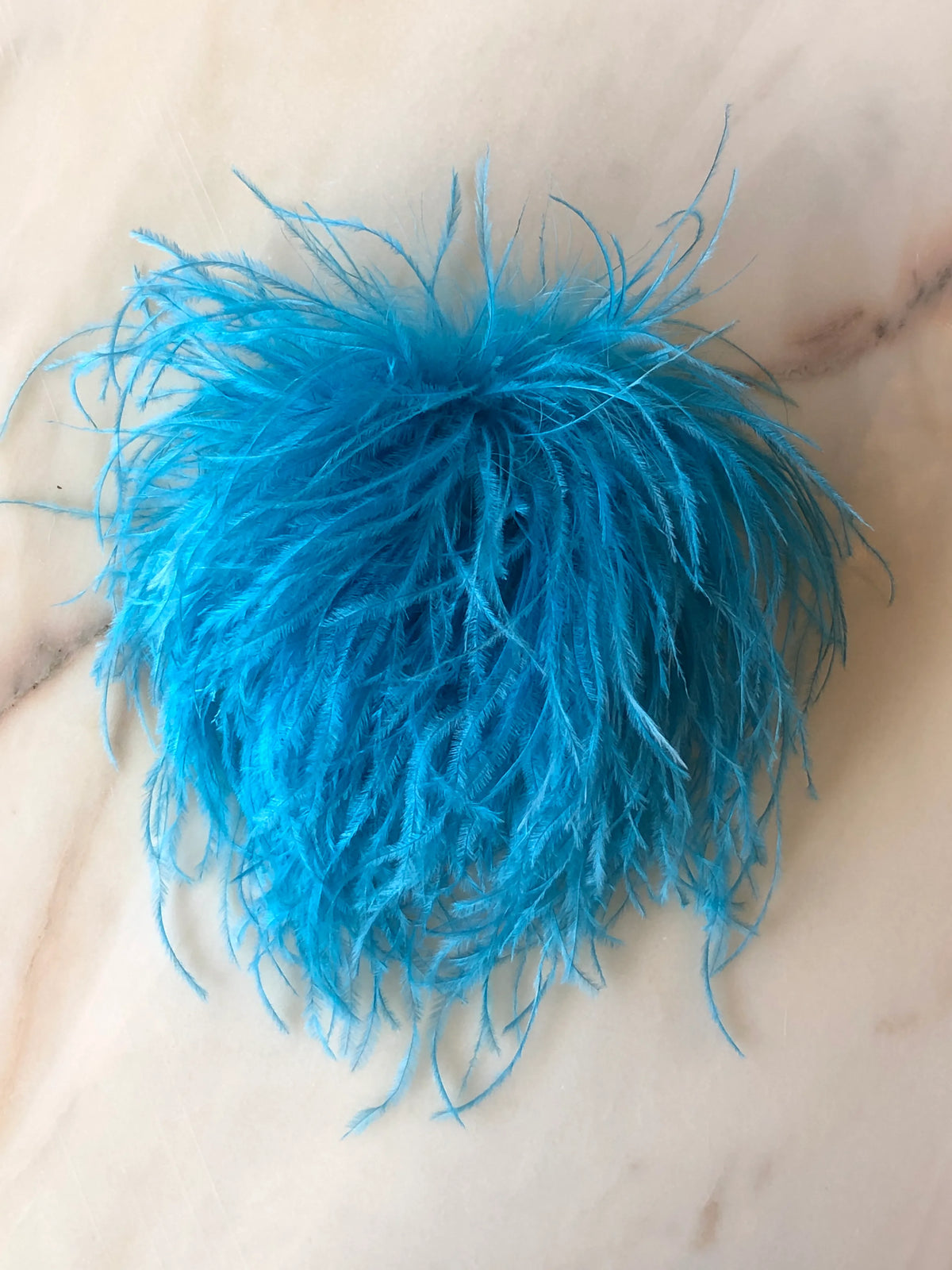 Broochella Le Pouffe Feather Brooch in vibrant blue, featuring a voluminous arrangement of soft, wispy feathers, photographed on a pale white marble background with subtle grey and blush veining.