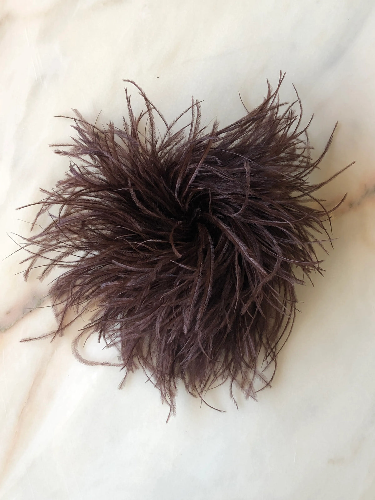 Broochella Le Pouffe Feather Brooch in rich chocolate brown, featuring a voluminous arrangement of soft, wispy ostrich feathers, photographed on a pale white marble background with subtle grey and blush veining.