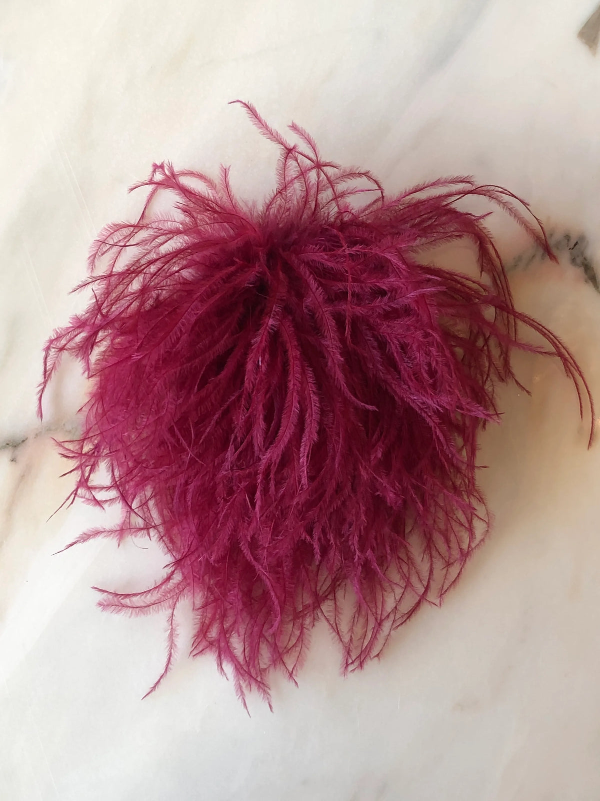 Broochella Le Pouffe Feather Brooch in rich burgundy, featuring a voluminous arrangement of soft, wispy feathers, photographed on a pale white marble background with subtle grey and blush veining.