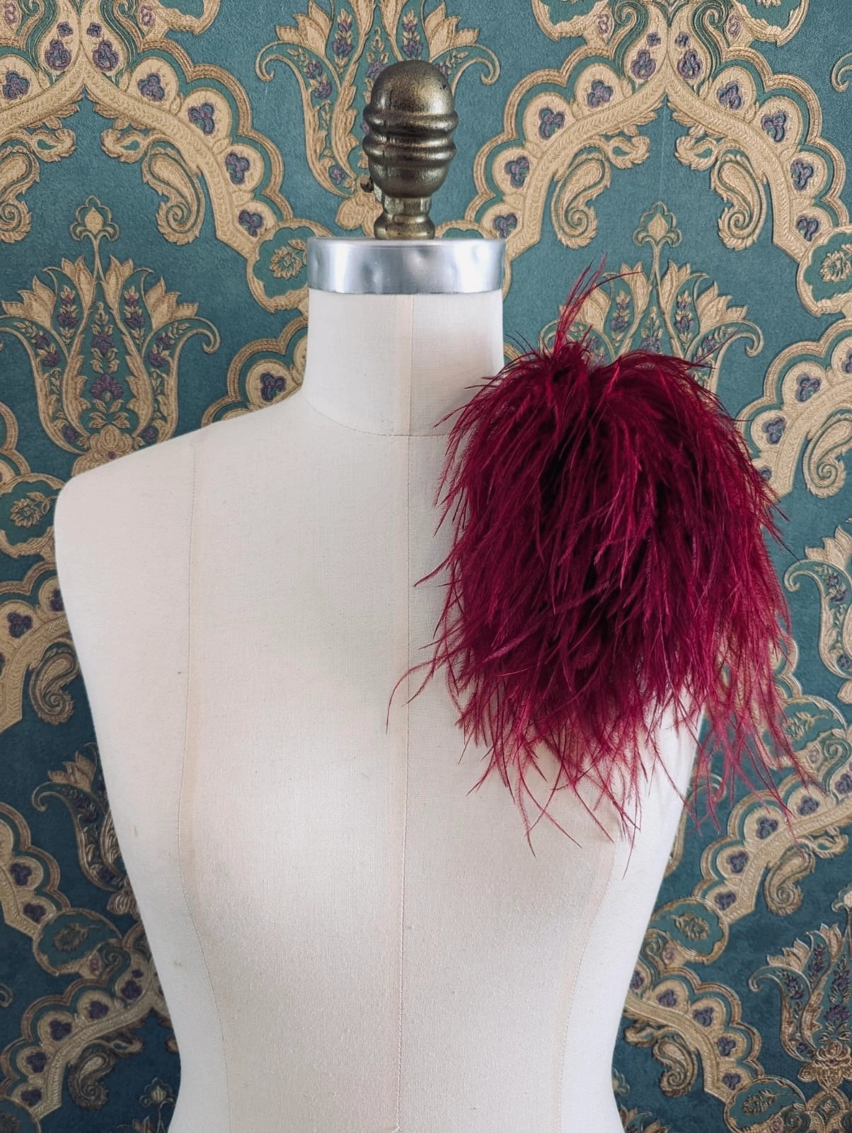 Broochella le pouffe handmade designer feather brooch in burgundy.  It is pinned to a mannequin. The background is green and gold damask wallpaper