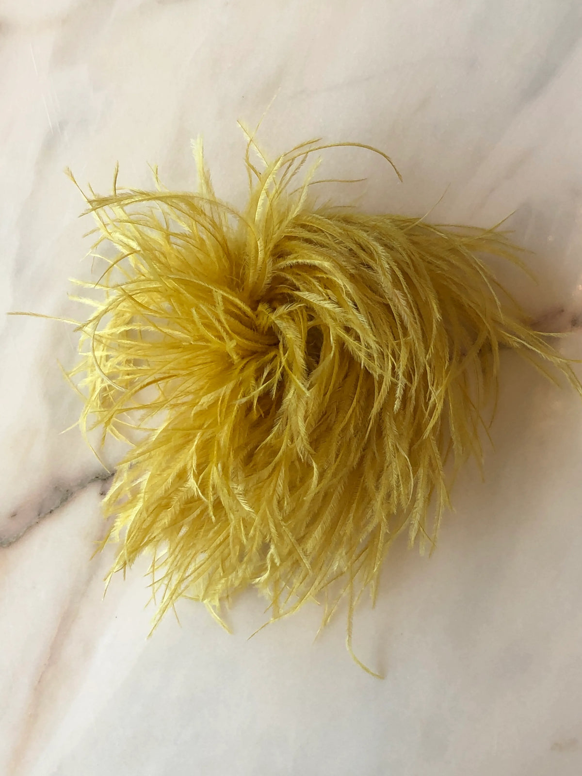Broochella Le Pouffe Feather Brooch in gold, featuring a voluminous arrangement of soft, wispy feathers, photographed on a pale white marble background with subtle grey and blush veining.