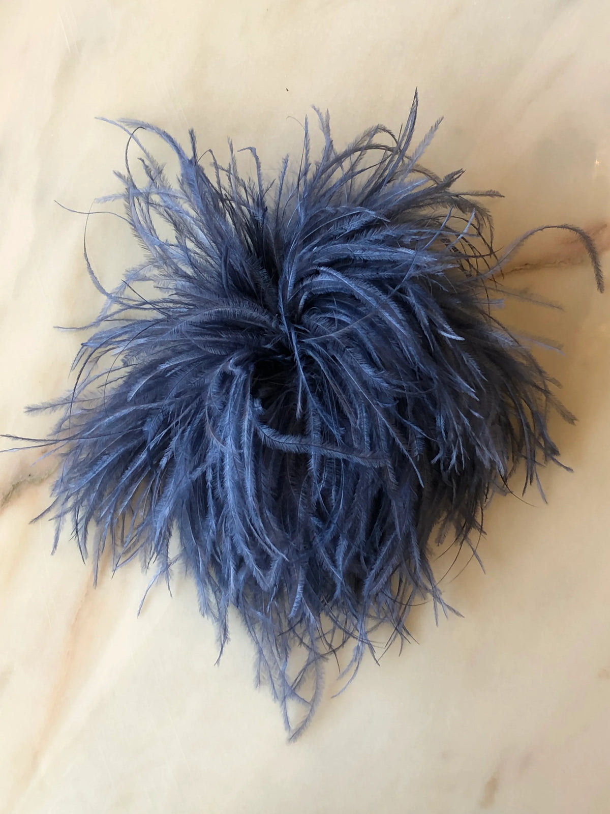 Broochella Le Pouffe Feather Brooch in grey, featuring a voluminous arrangement of soft, wispy feathers, photographed on a pale white marble background with subtle grey and blush veining.