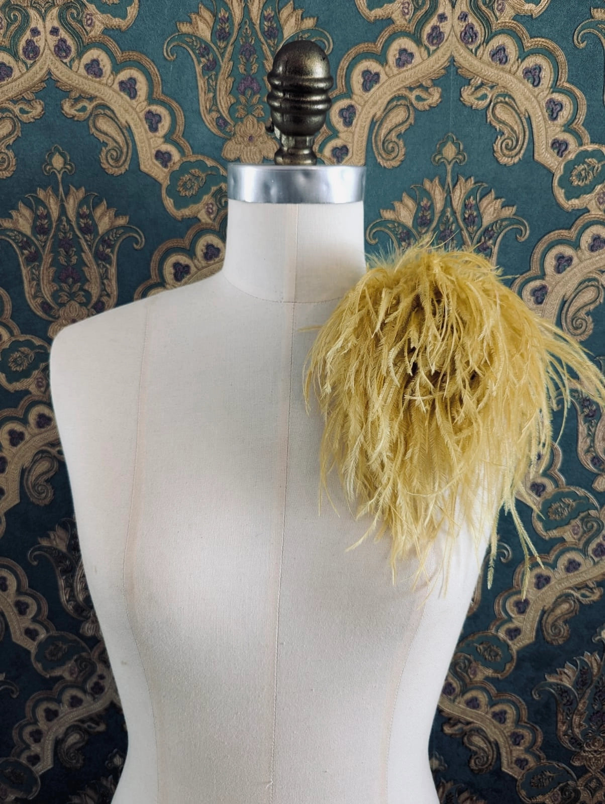 Broochella le pouffe handmade designer feather brooch in gold.  It is pinned to a mannequin. The background is green and gold damask wallpaper