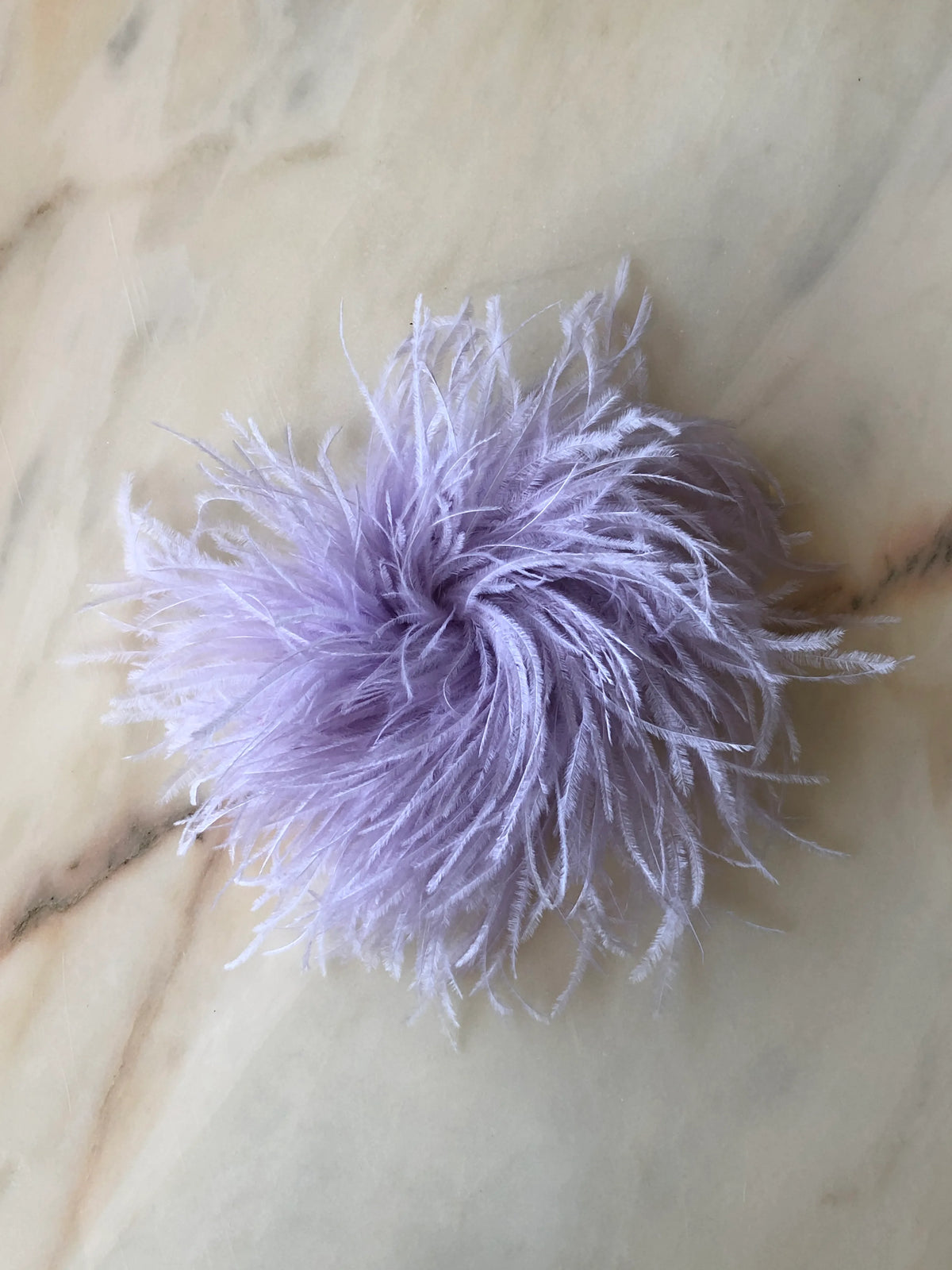 Broochella Le Pouffe Feather Brooch in soft lilac, featuring a voluminous arrangement of wispy ostrich feathers, photographed on a pale white marble background with subtle grey and blush veining.