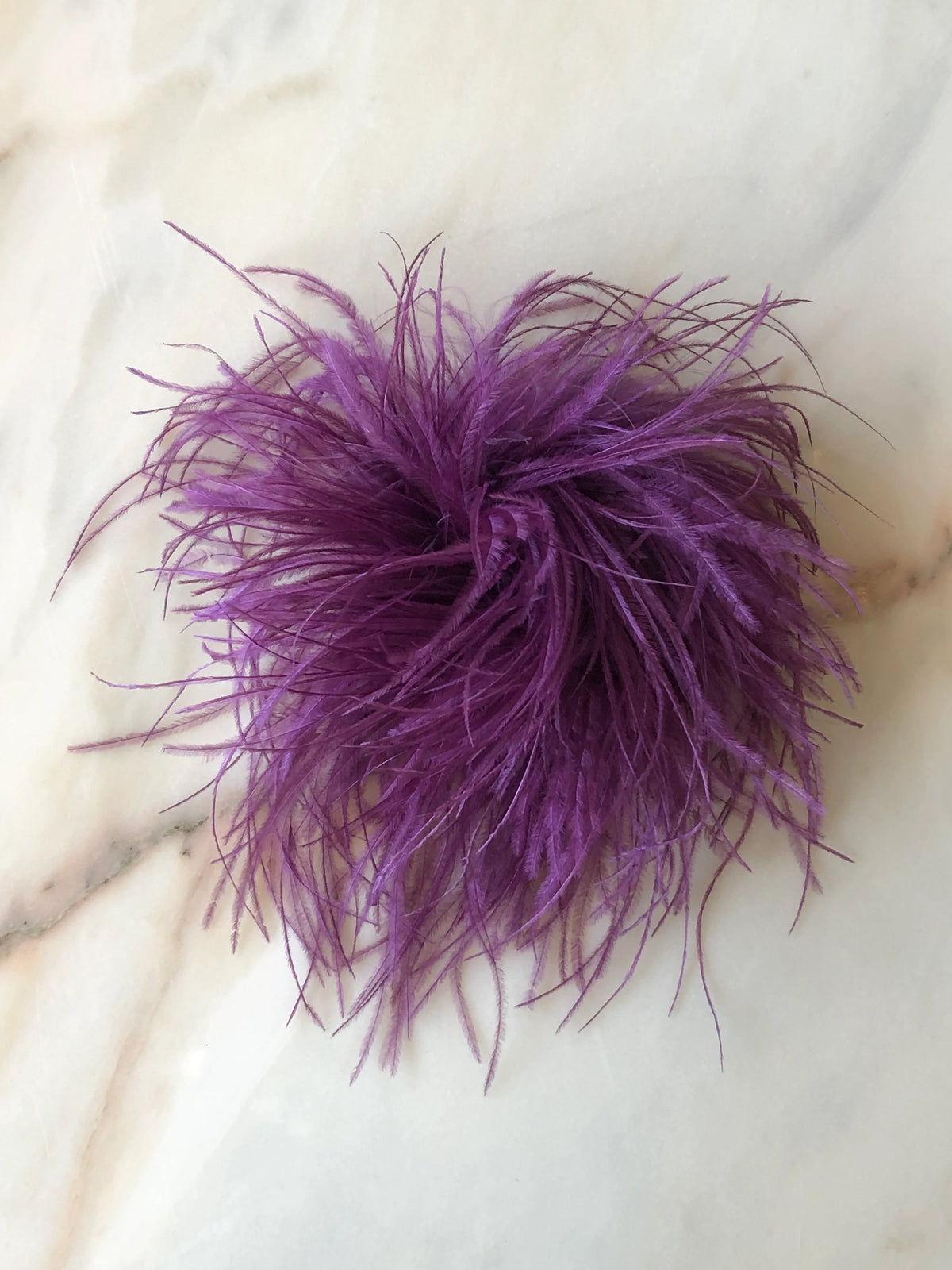 Broochella Le Pouffe Feather Brooch in rich magenta, featuring a voluminous arrangement of soft, wispy ostrich feathers, photographed on a pale white marble background with subtle grey and blush veining.