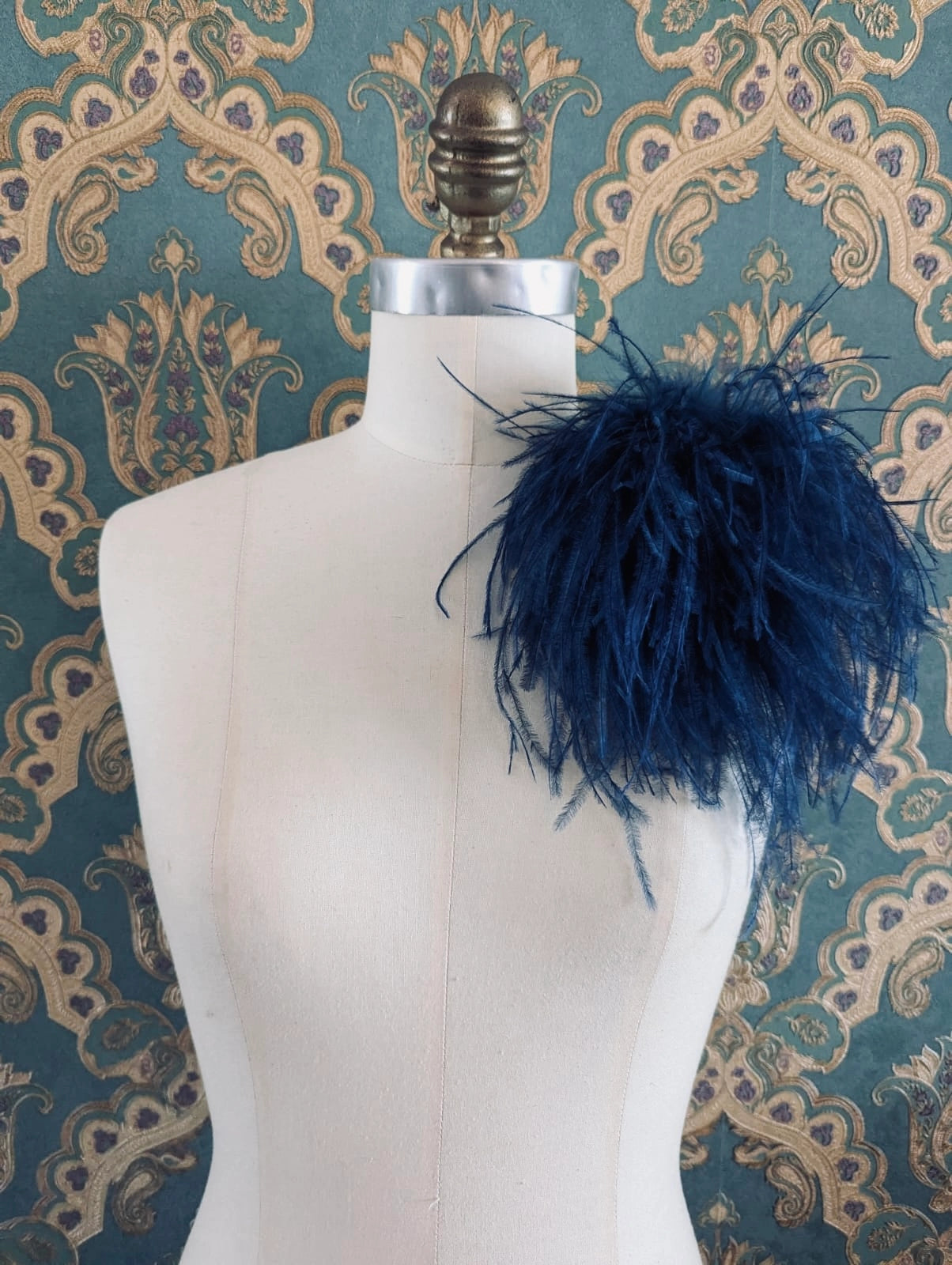 Broochella le pouffe handmade designer feather brooch in navy.  It is pinned to a mannequin. The background is green and gold damask wallpaper