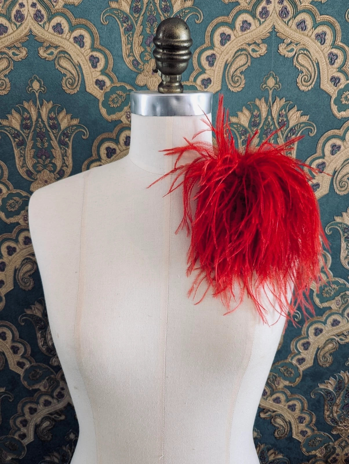 Broochella le pouffe handmade designer feather brooch in red.  It is pinned to a mannequin. The background is green and gold damask wallpaper