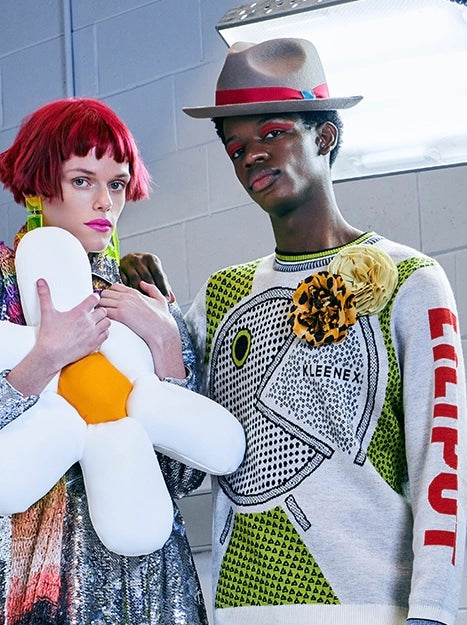 Two models posing in streetwear fashion. The model on the left has bright red hair and holds a large white daisy plush toy with an orange centre. He is wearing the Broochella Leopard flower brooch and the yellow Pom Pon brooch. 
