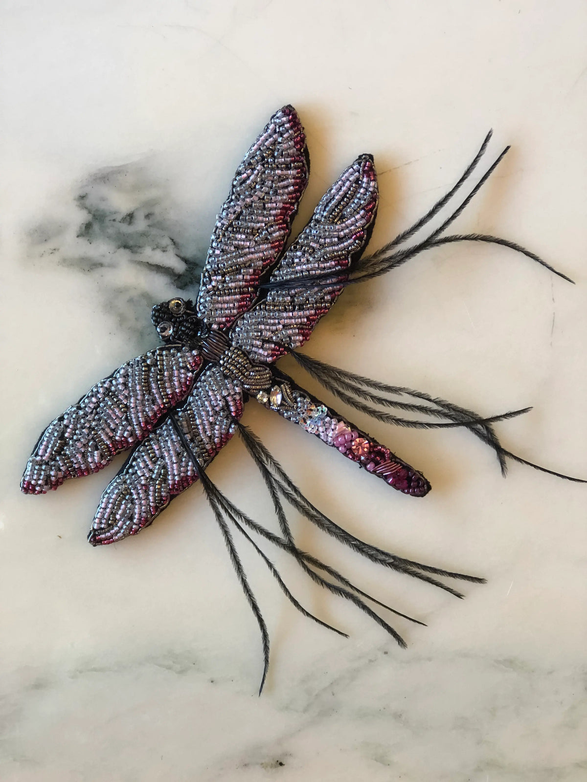 Broochella Libellula Dragonfly Hand-Beaded Brooch featuring intricate silver, lilac and pink beadwork, four embellished wings, crystal detailing and delicate black feather accents on a cream marble background.