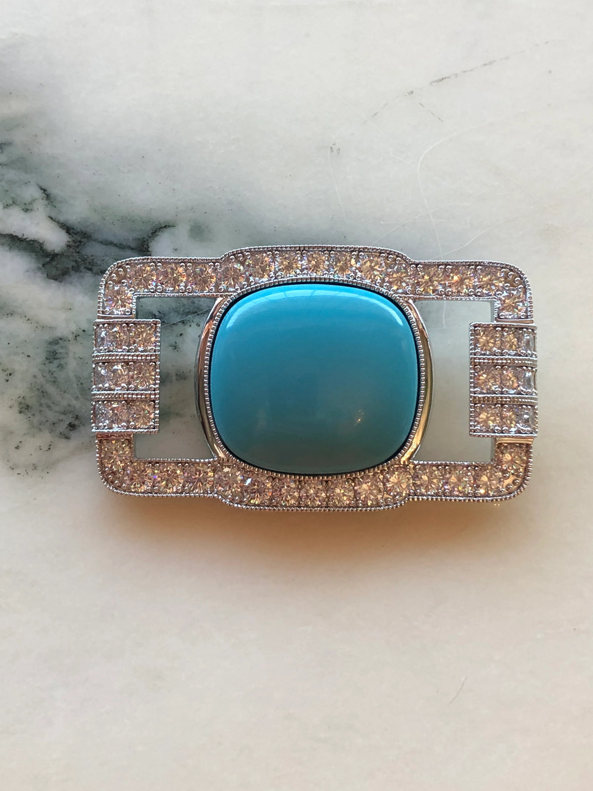 Broochella Libertenè Brooch Pin featuring a vibrant turquoise blue cabochon centre framed by sparkling clear crystals in a polished silver-tone geometric setting on a cream marble background.