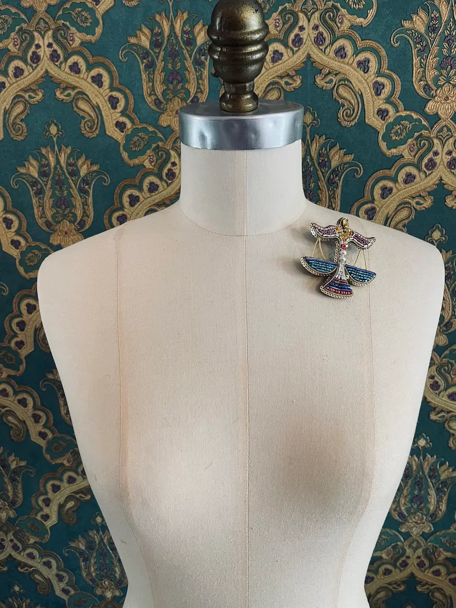 Broochella libra hand beaded zodiac designer brooch. Pictured on a mannequin. The background is green and gold damask wallpaper