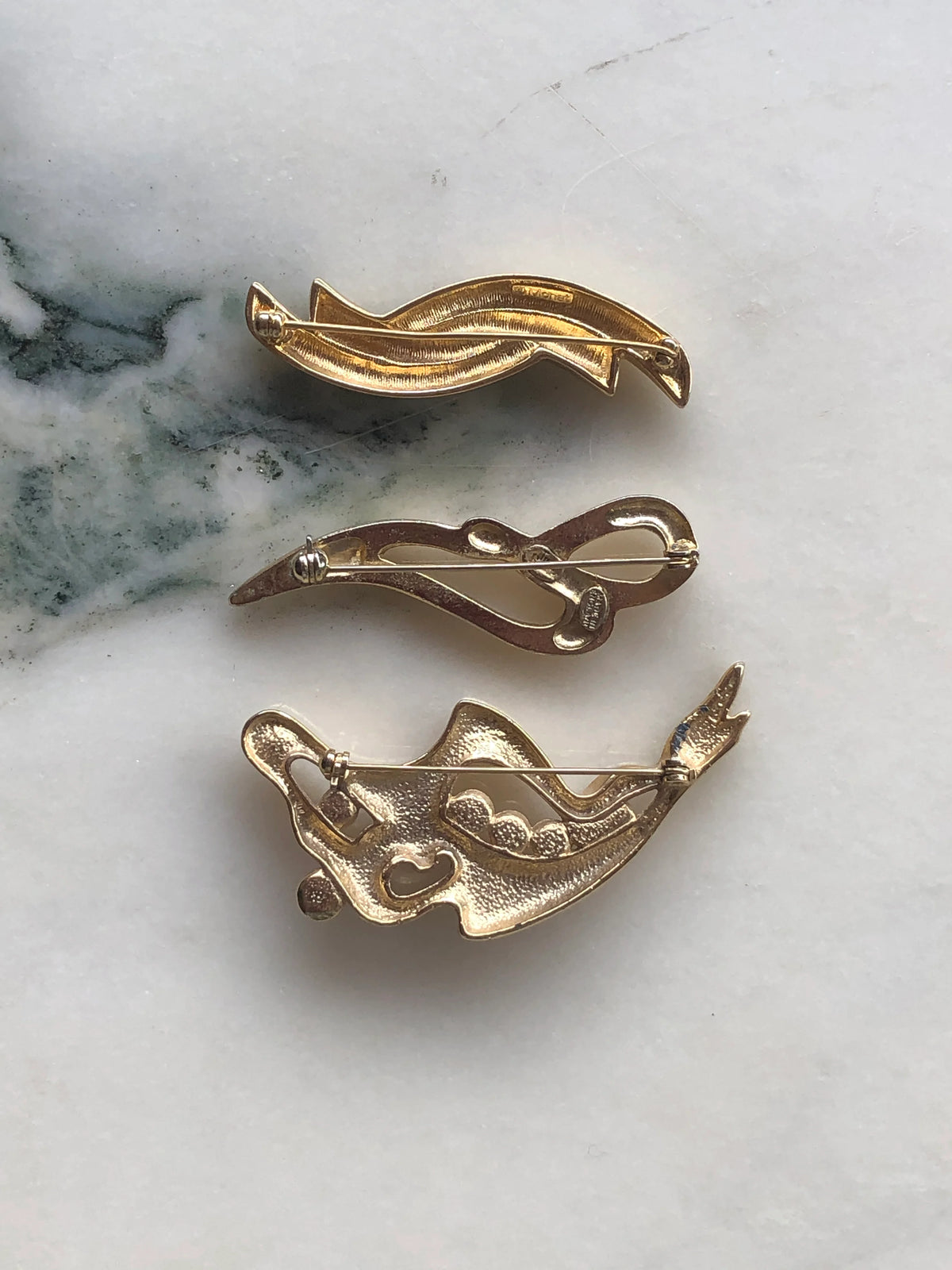Broochella Linea Lucente vintage brooch set back view showing the gold-tone metalwork, vintage markings and pin fastenings of all three brooches on a white and green veined marble background.