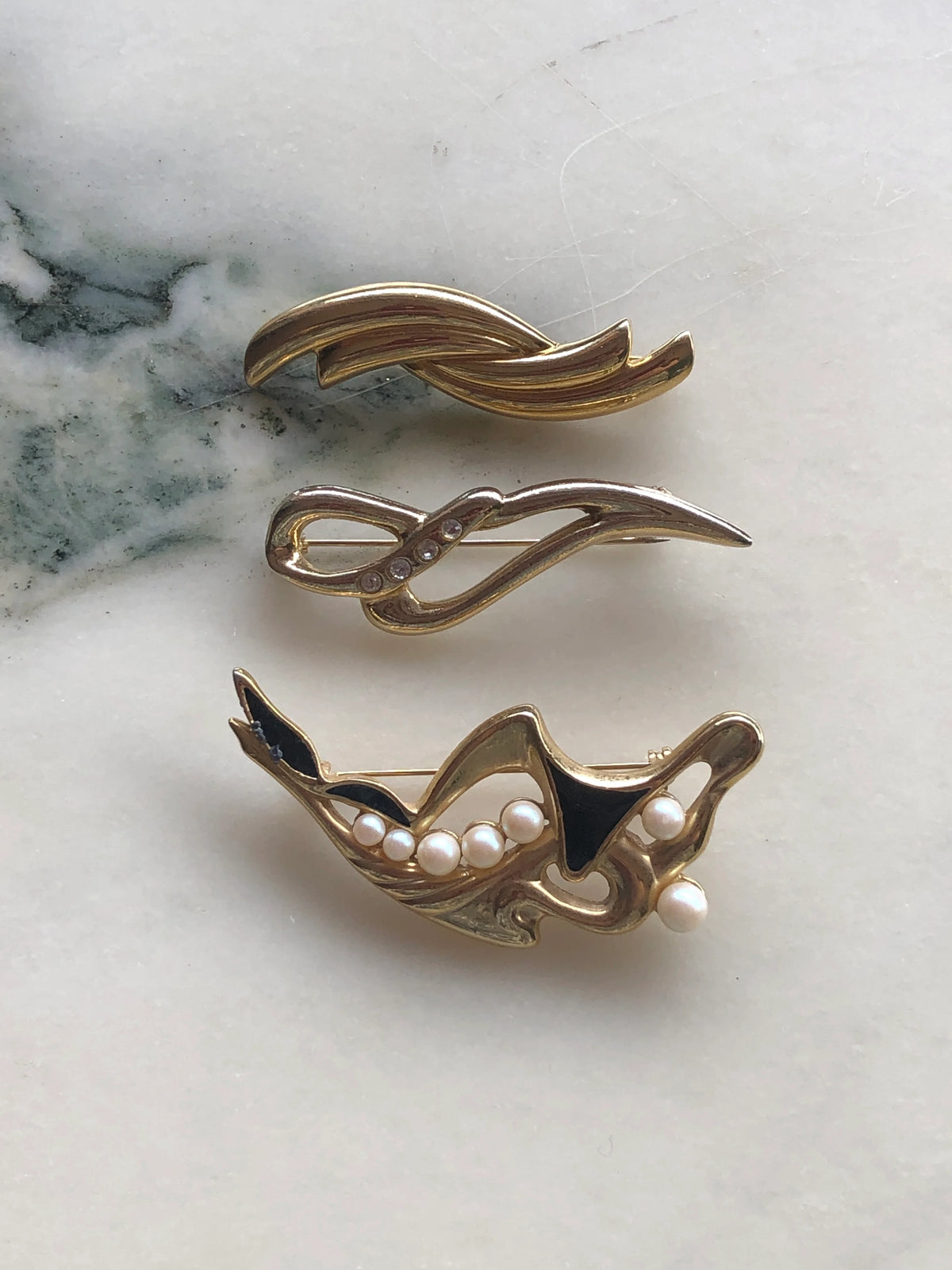 Broochella Linea Lucente vintage brooch set front view featuring three abstract gold-tone designs with crystal, black enamel and pearl details, photographed on a white and green veined marble background.