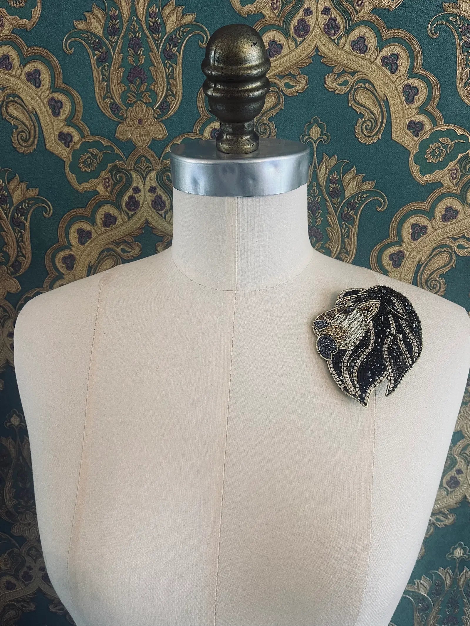 Broochella black and gold hand beaded large lion brooch. Pictured on a mannequin. The background is green and gold damask wallpaper