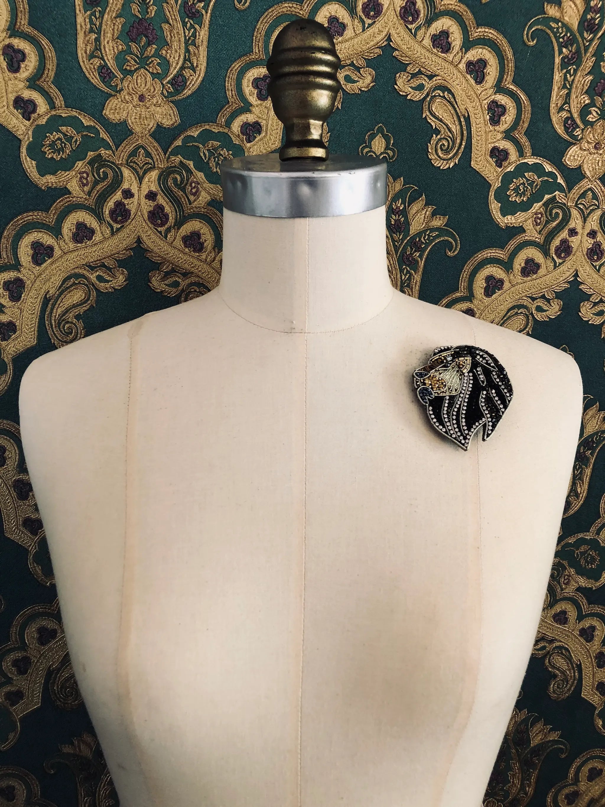 Broochella hand beaded black and goild small lion brooch. Pictured on a mannequin. The background is green and gold damask wallpaper