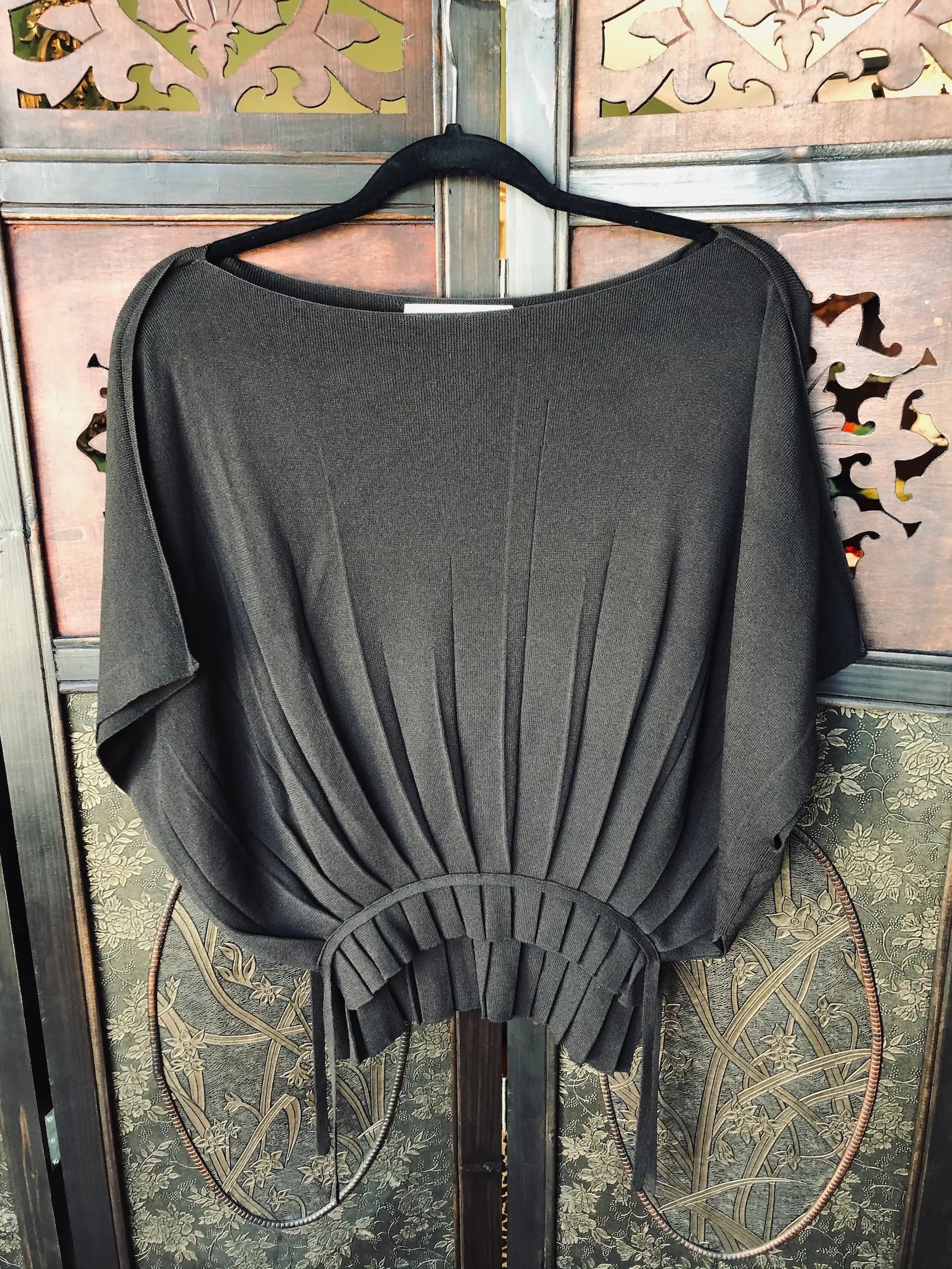 Broochella Livinia Conti black knit top with pleated waist detailing. It is hung against an ornate, dark wood room divider. 