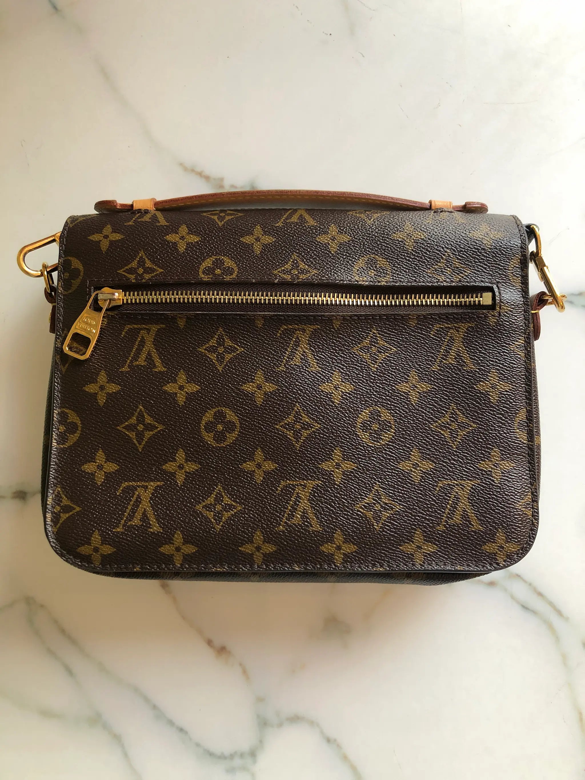 The back view of A Broochella Louis Vuitton Pochette Metis monogram shoulder bag in brown coated canvas with gold-tone hardware, resting on a white marble surface. The bag features the classic LV monogram pattern, a top flap with a gold-tone padlock closure, and an adjustable black leather shoulder strap. The background is white marble
