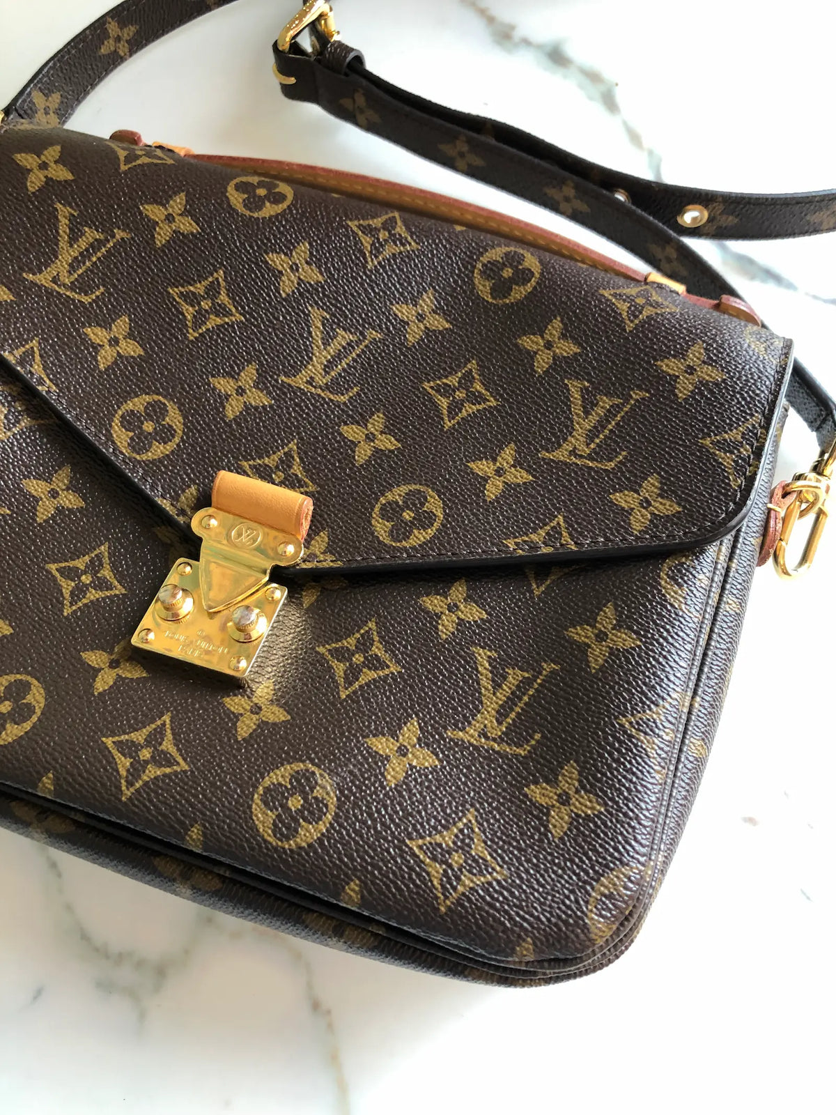 A Broochella Louis Vuitton Pochette Metis monogram shoulder bag in brown coated canvas with gold-tone hardware, resting on a white marble surface. The bag features the classic LV monogram pattern, a top flap with a gold-tone padlock closure, and an adjustable black leather shoulder strap. The background is white marble