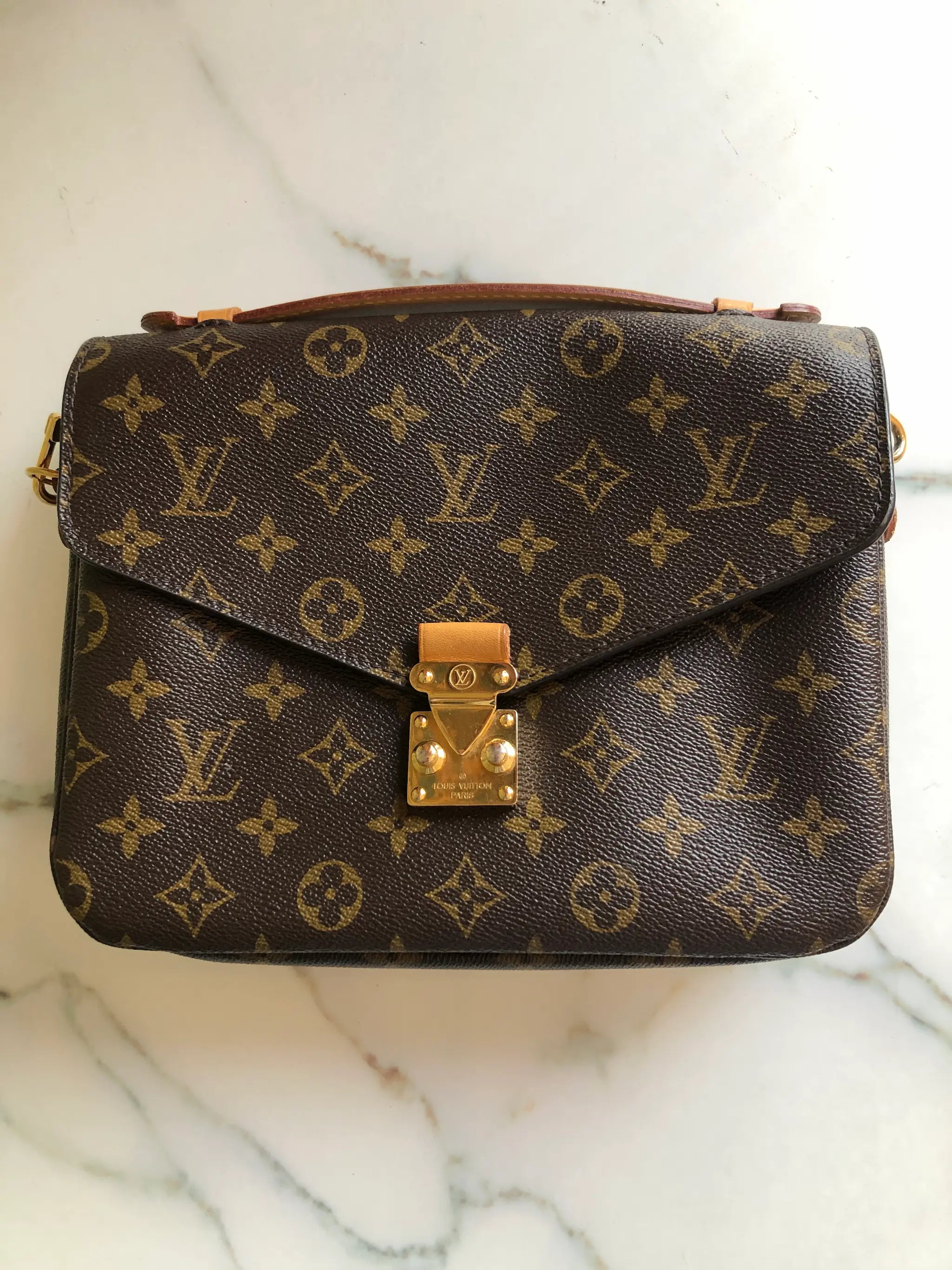 A Broochella Louis Vuitton Pochette Metis monogram shoulder bag in brown coated canvas with gold-tone hardware, resting on a white marble surface. The bag features the classic LV monogram pattern, a top flap with a gold-tone padlock closure, and an adjustable black leather shoulder strap. The background is white marble