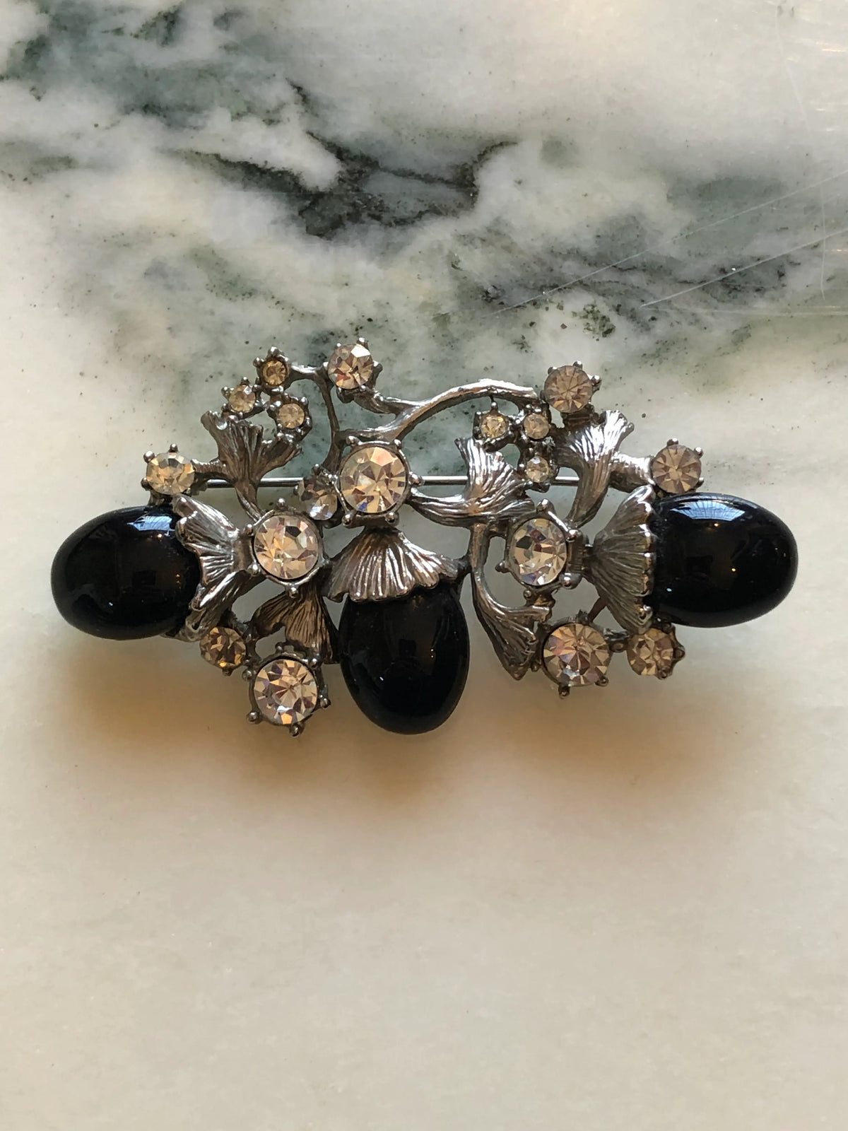 Broochella Luciana Vintage Black Cabochon Floral Brooch featuring three glossy black oval cabochons, clear crystals and sculptural silver-tone leaves on a cream and grey marble background.