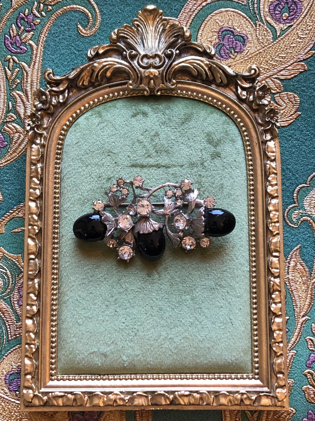 Broochella Luciana Vintage Black Cabochon Floral Brooch featuring three glossy black oval cabochons, clear crystals and sculptural silver-tone leaves. It is presented on a green velvet tray with an ornate gold frame. The background is green and gold damask wallpaper.