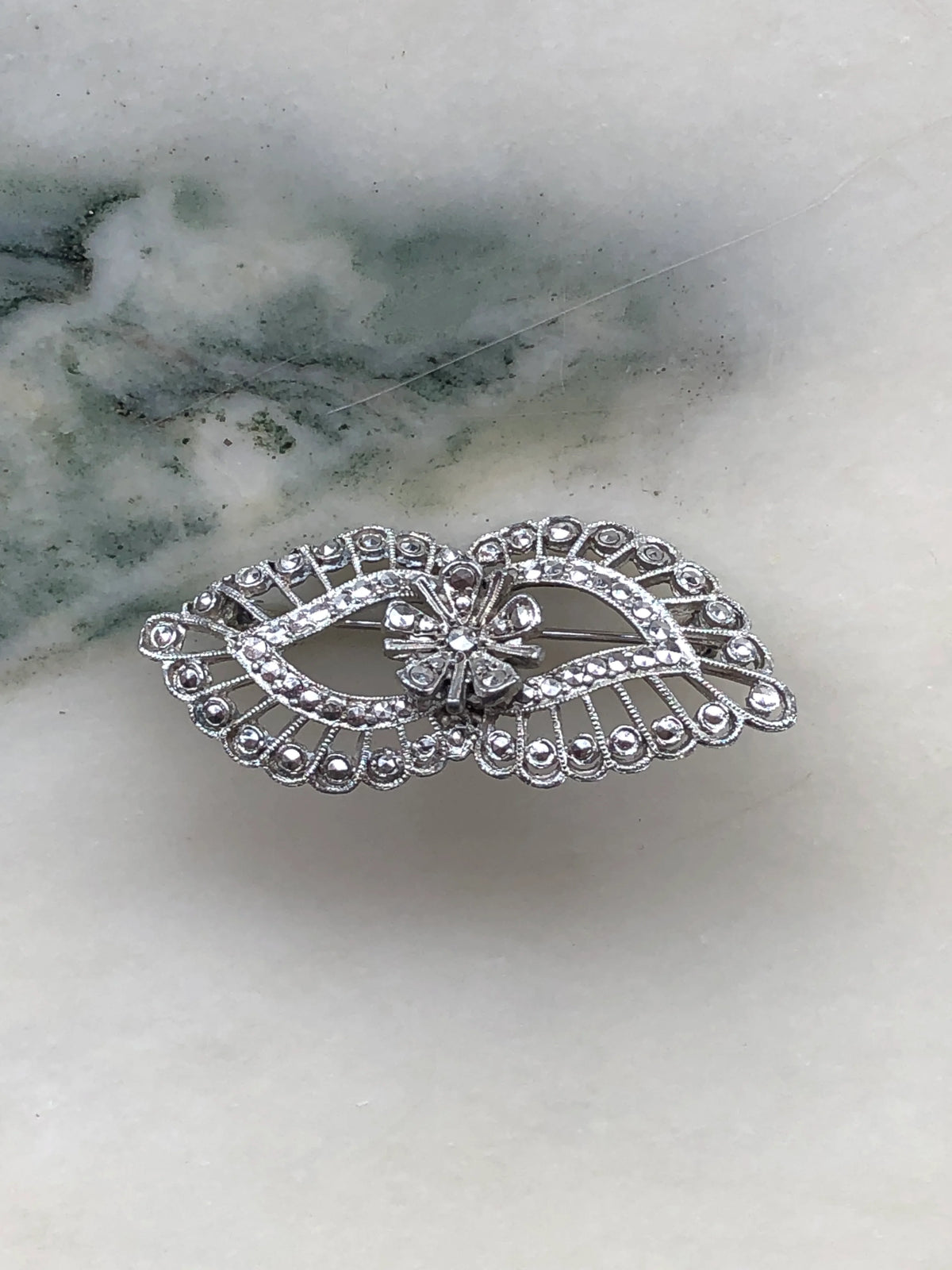 Broochella Luciana Vintage Silver Filigree Crystal Brooch in silver-tone metal, featuring an elongated oval silhouette, intricate openwork scroll detailing, sparkling clear crystals and a delicate central floral design, photographed on white marble with grey veining.