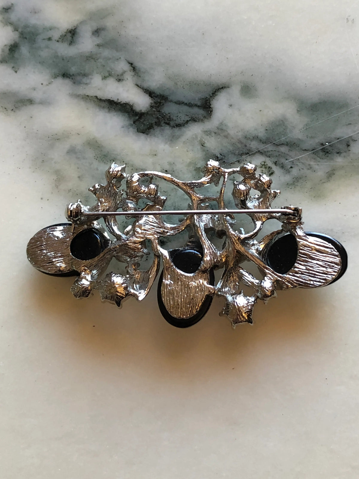 Broochella Lucilla Vintage Gunmetal Cabochon Floral Brooch featuring three polished gunmetal oval cabochons, clear crystals and sculptural silver-tone leaves on a cream and grey marble background.