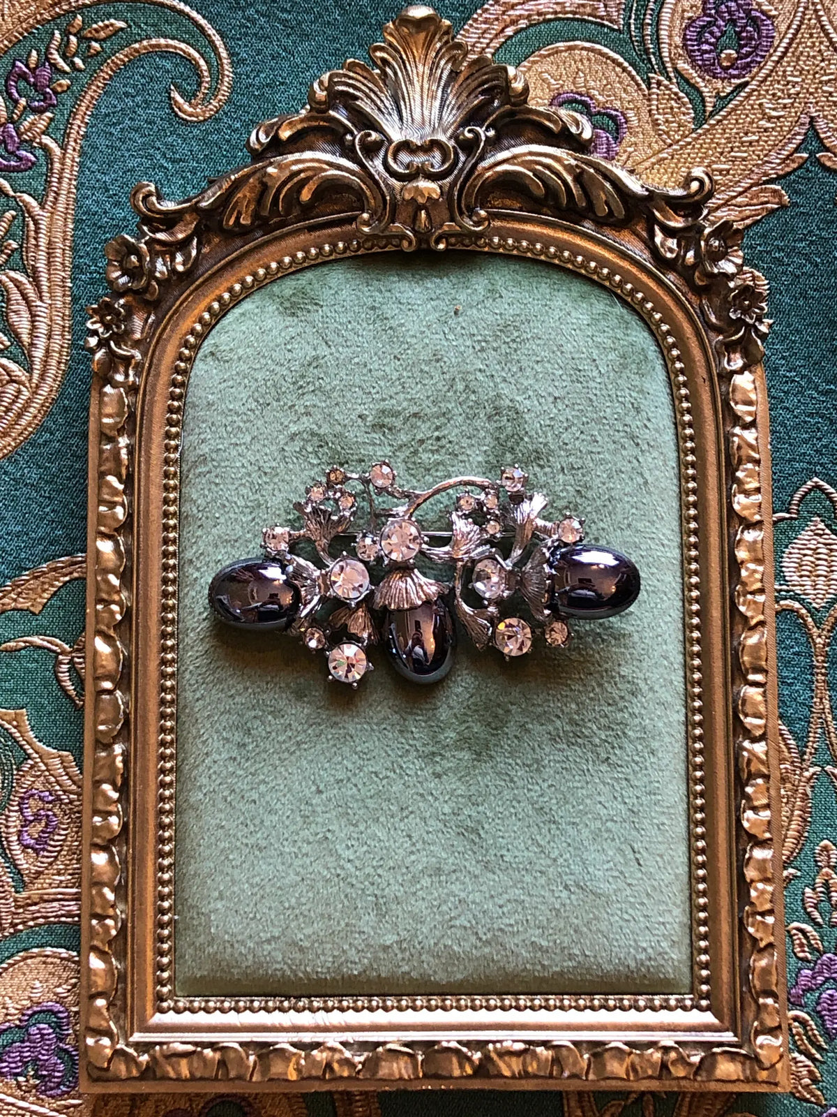 Broochella Lucilla Vintage Gunmetal Cabochon Floral Brooch featuring three polished gunmetal oval cabochons, clear crystals and sculptural silver-tone leaves. It is presented on a green velvet tray with an ornate gold frame. The background is green and gold damask wallpaper. 