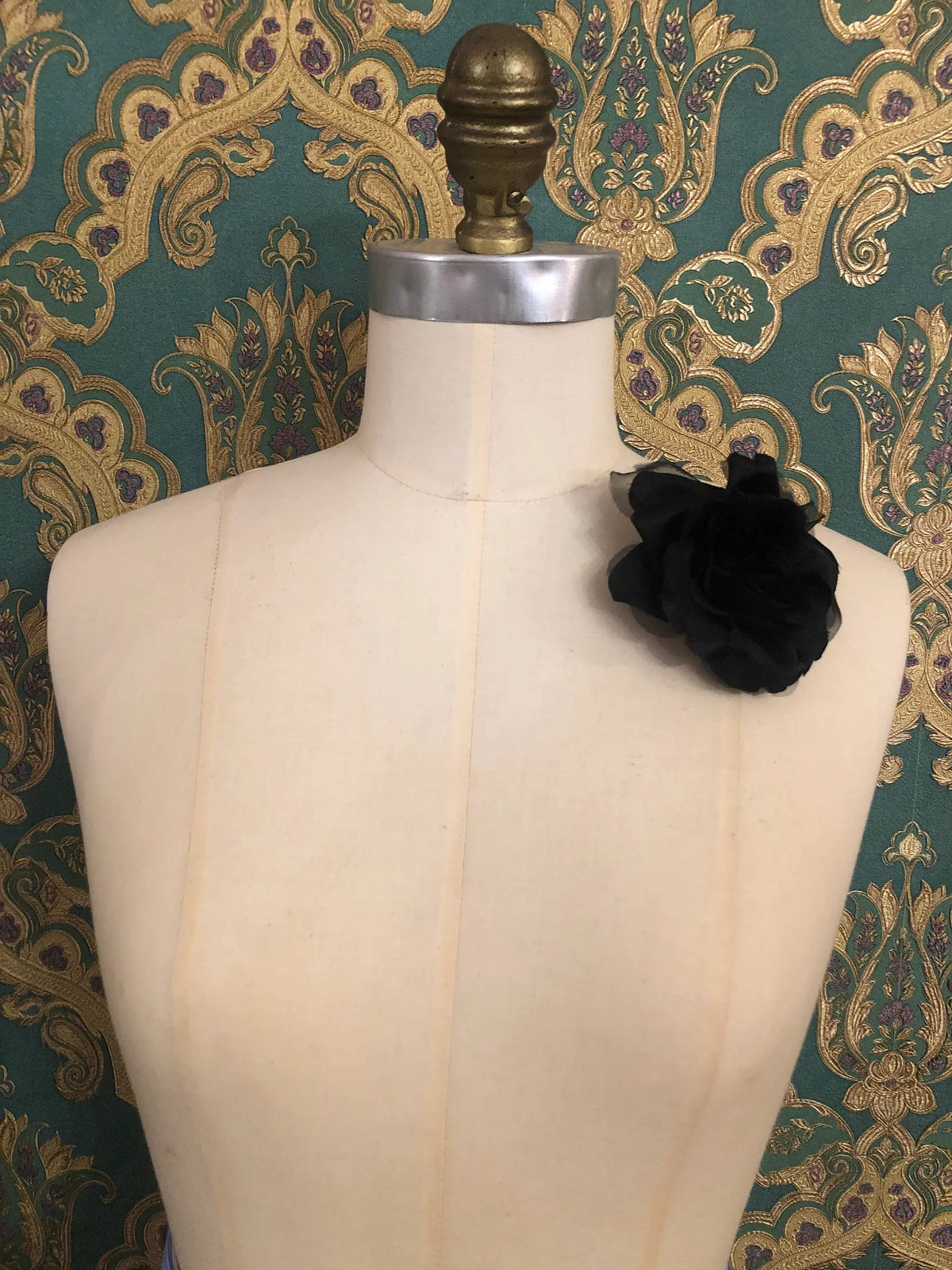 Broochella Ludovica black flower brooch pinned to a cream mannequin against green and gold damask wallpaper.