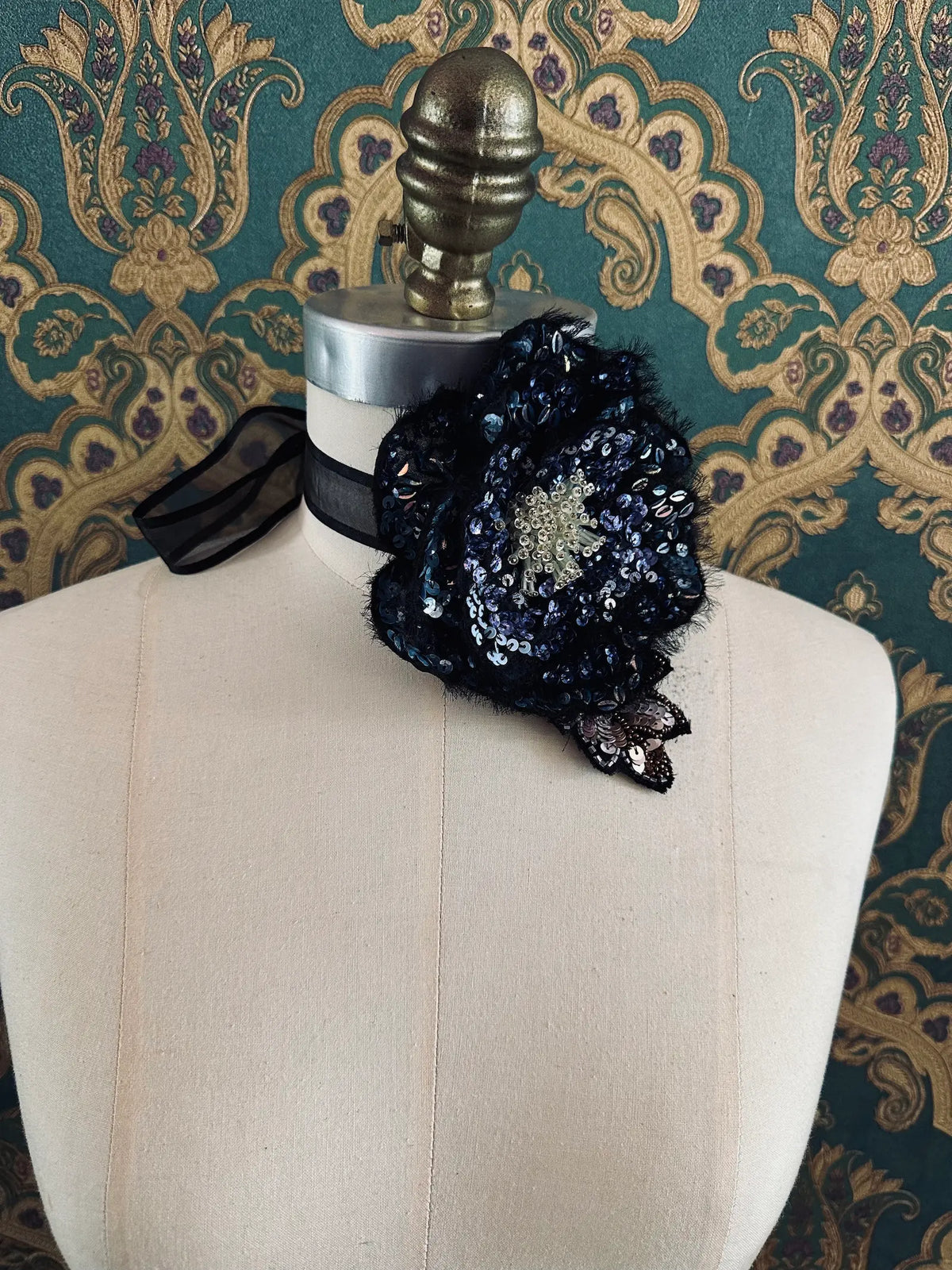 Broochella Luminosa hand beaded sequin choker and brooch in blue and black. Pictured on a mannequin. The background is green and gold damask wallpaper.