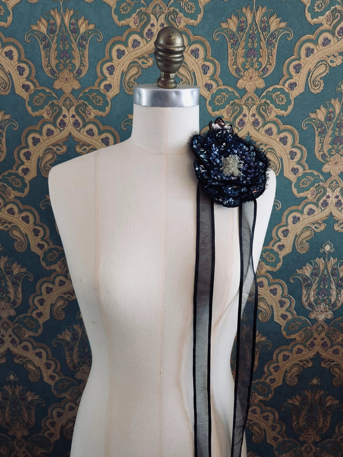 Broochella Luminosa hand beaded sequin choker and brooch in blue and black. Pictured on a mannequin. The background is green and gold damask wallpaper.