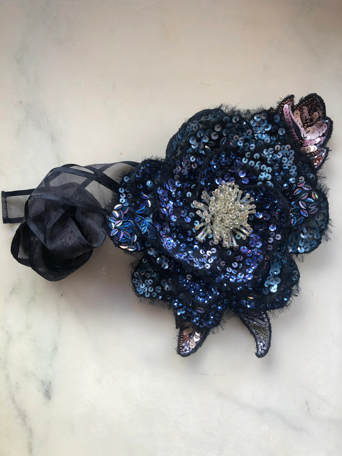 Broochella Luminosa large flower choker and brooch, hand embellished with navy and blue sequins, iridescent beads and a pearl-beaded centre, with black organza ribbon, photographed on white marble.