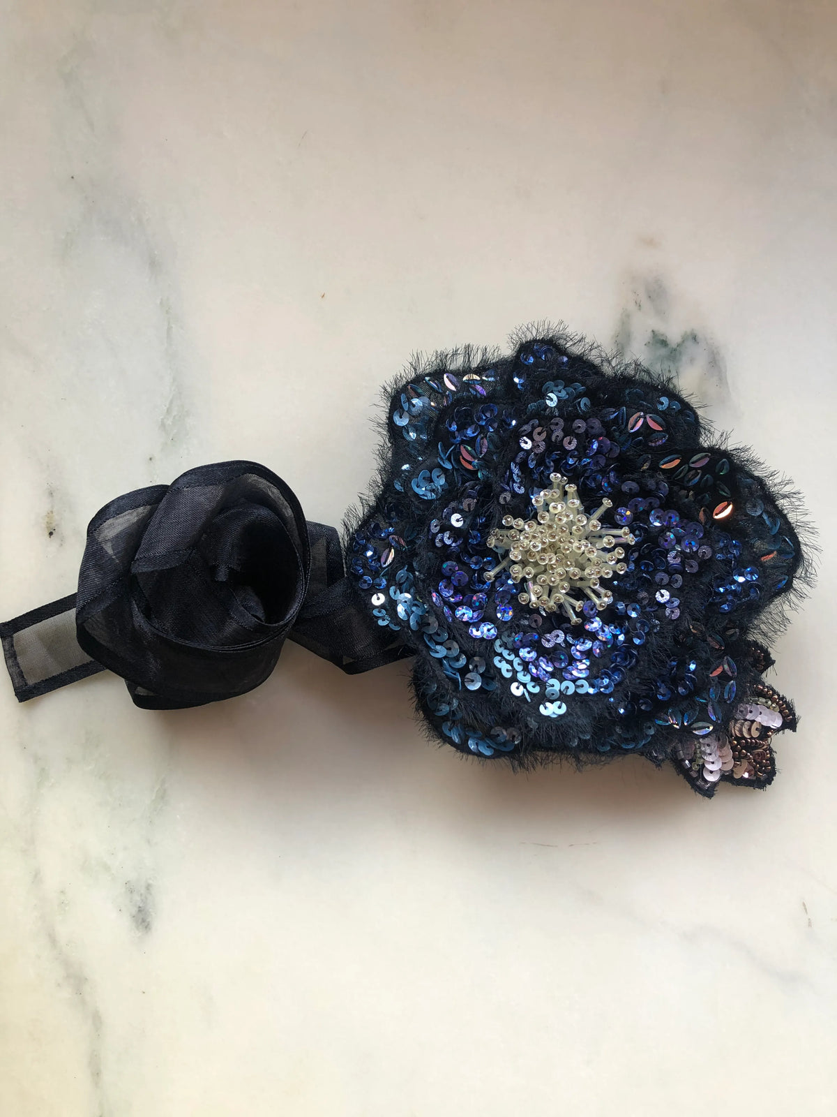 A Broochella Luminosa black and blue sequinned flower brooch with a black sheer organza rose and a silver rhinestone centre, displayed on a light marble surface.