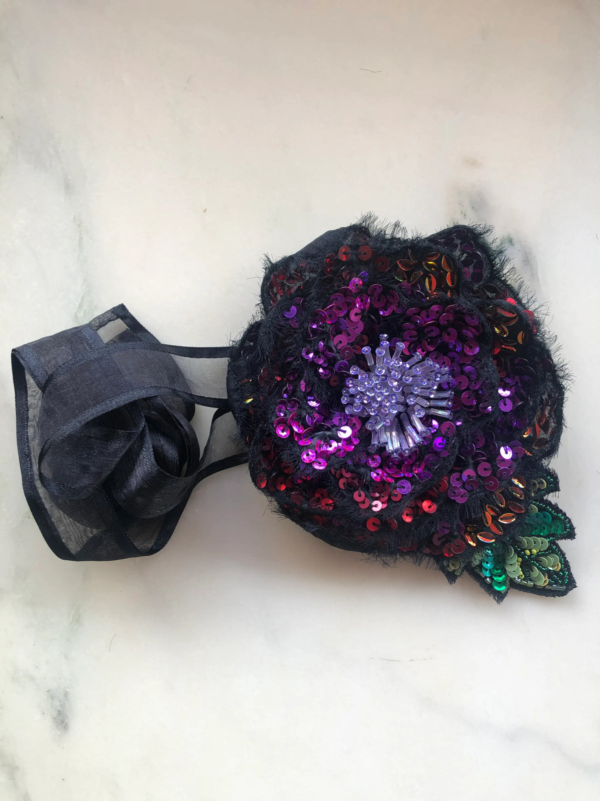 A Broochella Luminosa black, pink and red sequinned flower brooch with a black sheer organza rose and a silver rhinestone centre, displayed on a light marble surface.