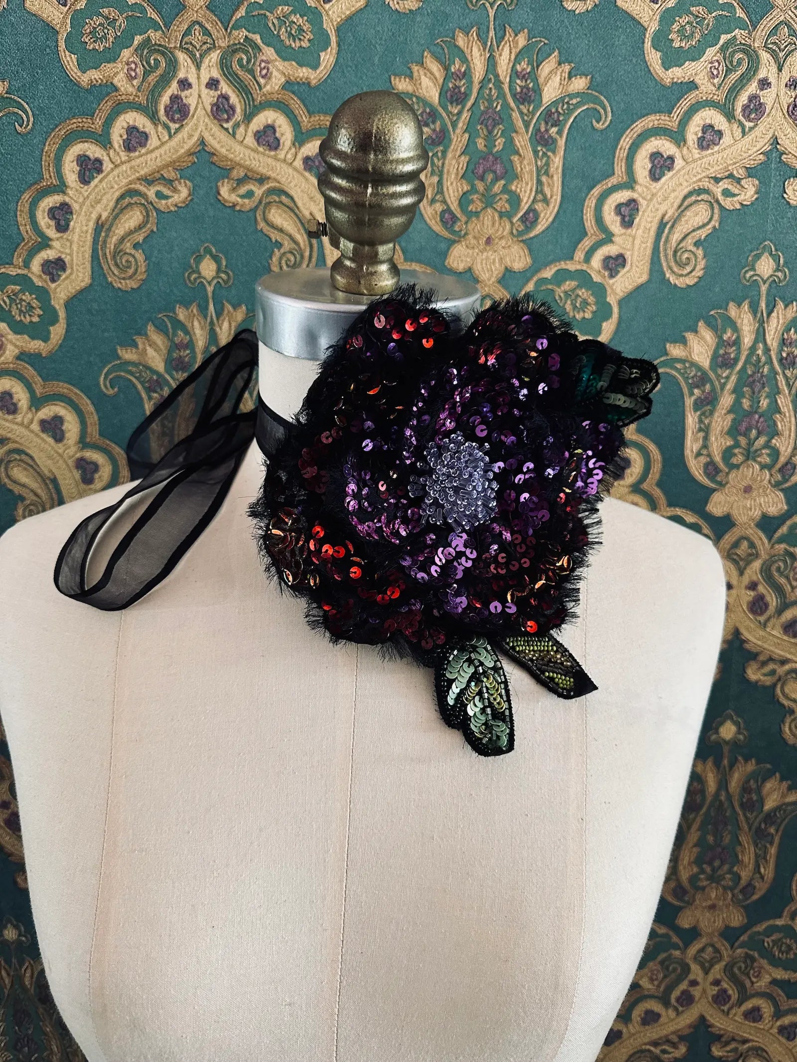 A Broochella luminosa  black, red and purple sequinned floral choker and brooch, with lace leaves and a sheer black ribbon bow, displayed on a cream-coloured dress form against a green and gold patterned wallpaper background.
