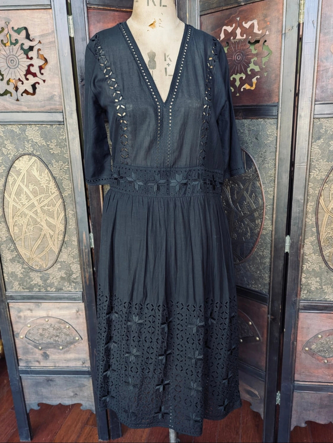 Broochella designer  Magali Pascal Black Midi Dress with broderie lace detailing, a v neck and 3/4 sleeves The dress is presented on a mannequin. The background is a dark wood room divider