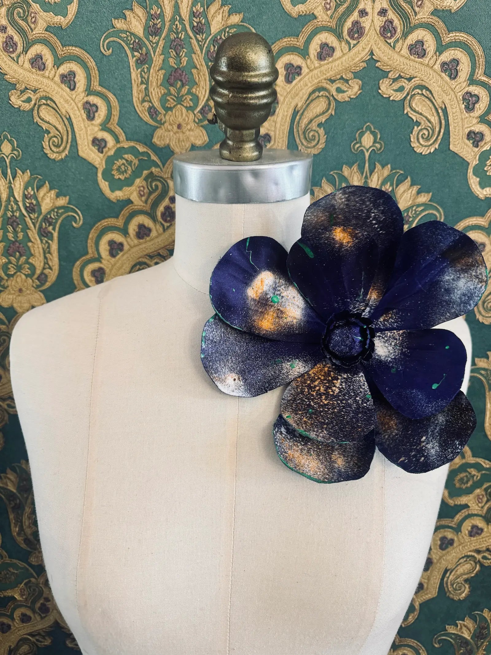 Broochella IMagnolia d'arte large blue flower brooch with multi coloured paint splatter. One of a kind customized piece. Pictured on a mannequin.  The background is green and gold damask wallpaper.