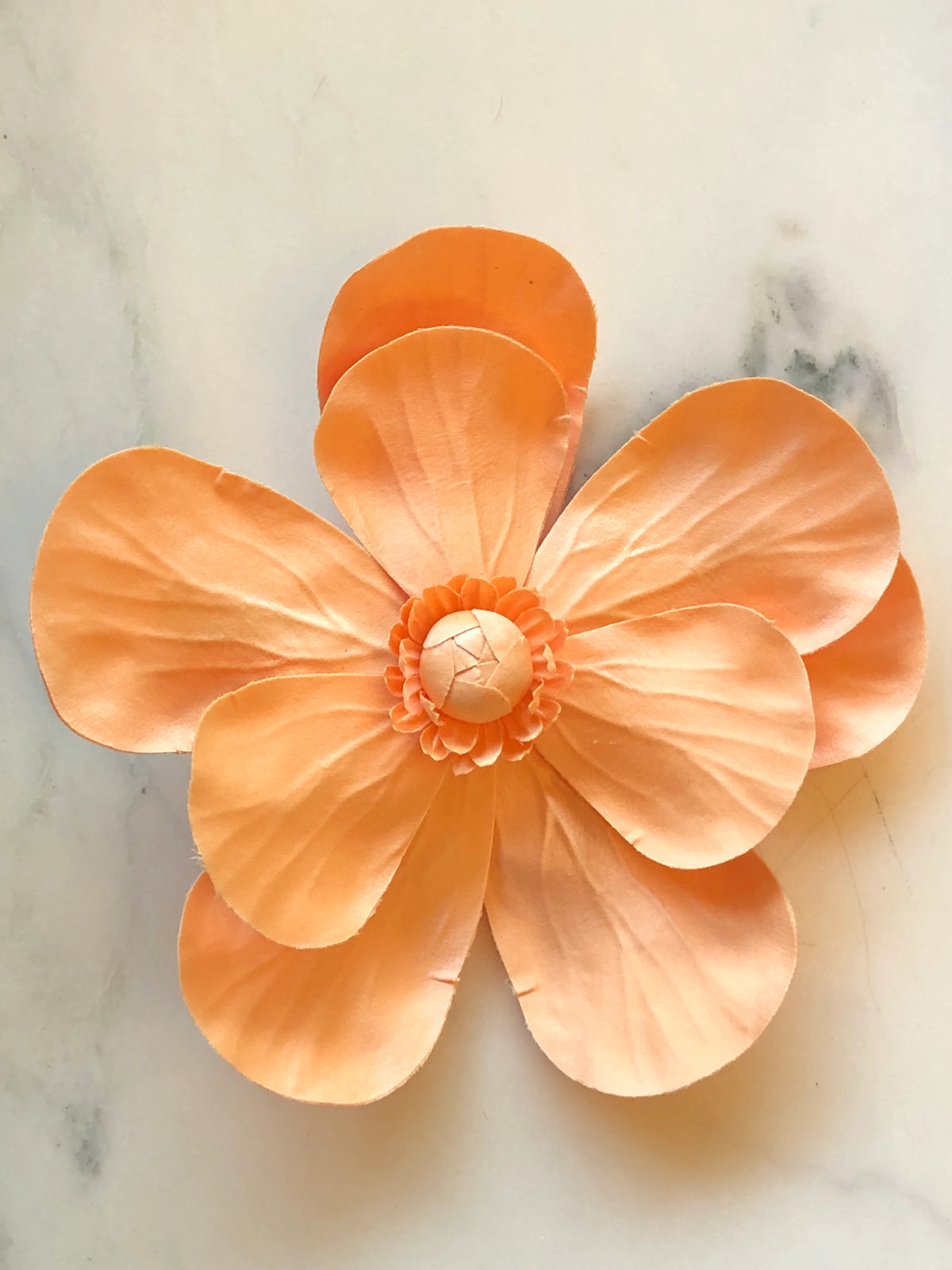 Broochella Magnolia Flower Brooch in warm orange, featuring oversized layered fabric petals with sculptural veining and a handcrafted floral centre on a cream marble background.