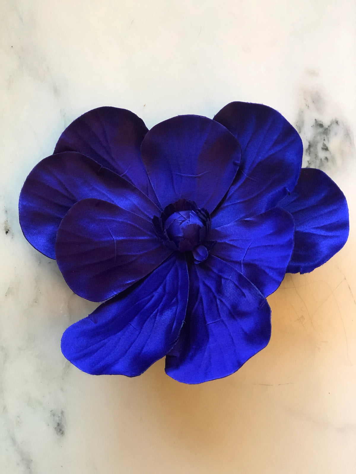 Broochella Magnolia Flower Brooch in rich purple, featuring oversized layered fabric petals with sculptural veining and a tonal handcrafted floral centre on a cream marble background.