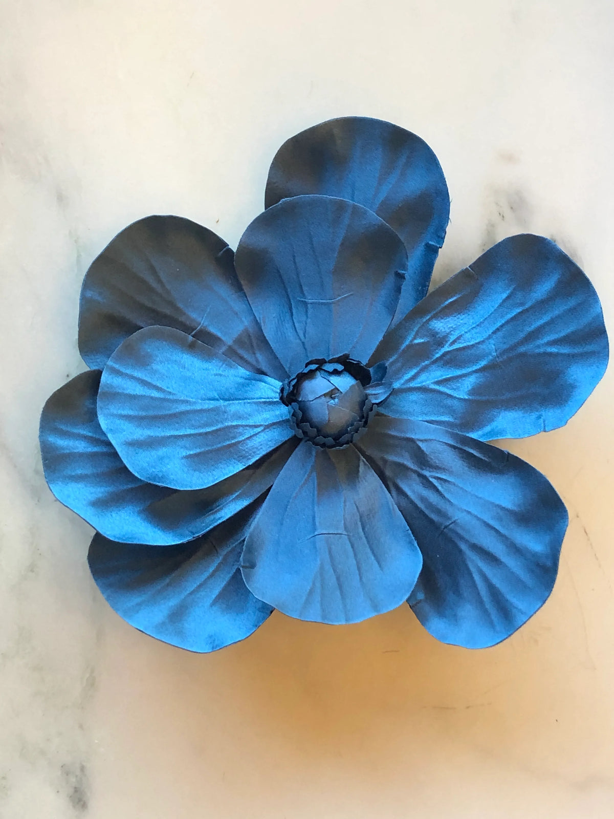 Broochella Magnolia Flower Brooch in teal blue, featuring oversized layered fabric petals with sculptural veining and a tonal handcrafted floral centre on a cream marble background.